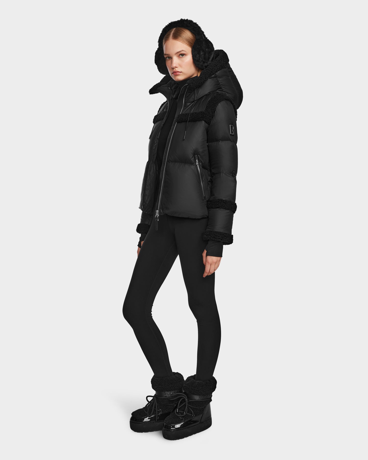 A person wearing the Rudsak JULIANA - L124509 Black/Black women’s short down puffer with RDS-certified down fill, fitted leggings, and winter boots stands against a plain white background.