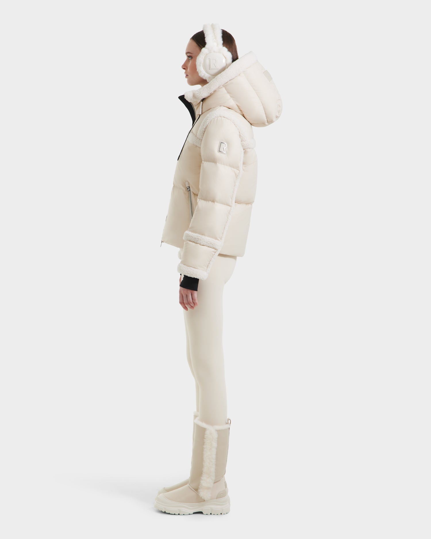 A woman in profile wears the Rudsak JULIANA - L124509 Vanilla short down puffer jacket with a hood, paired with matching leggings, earmuffs, and winter boots—an essential winter look—set against a plain white background.