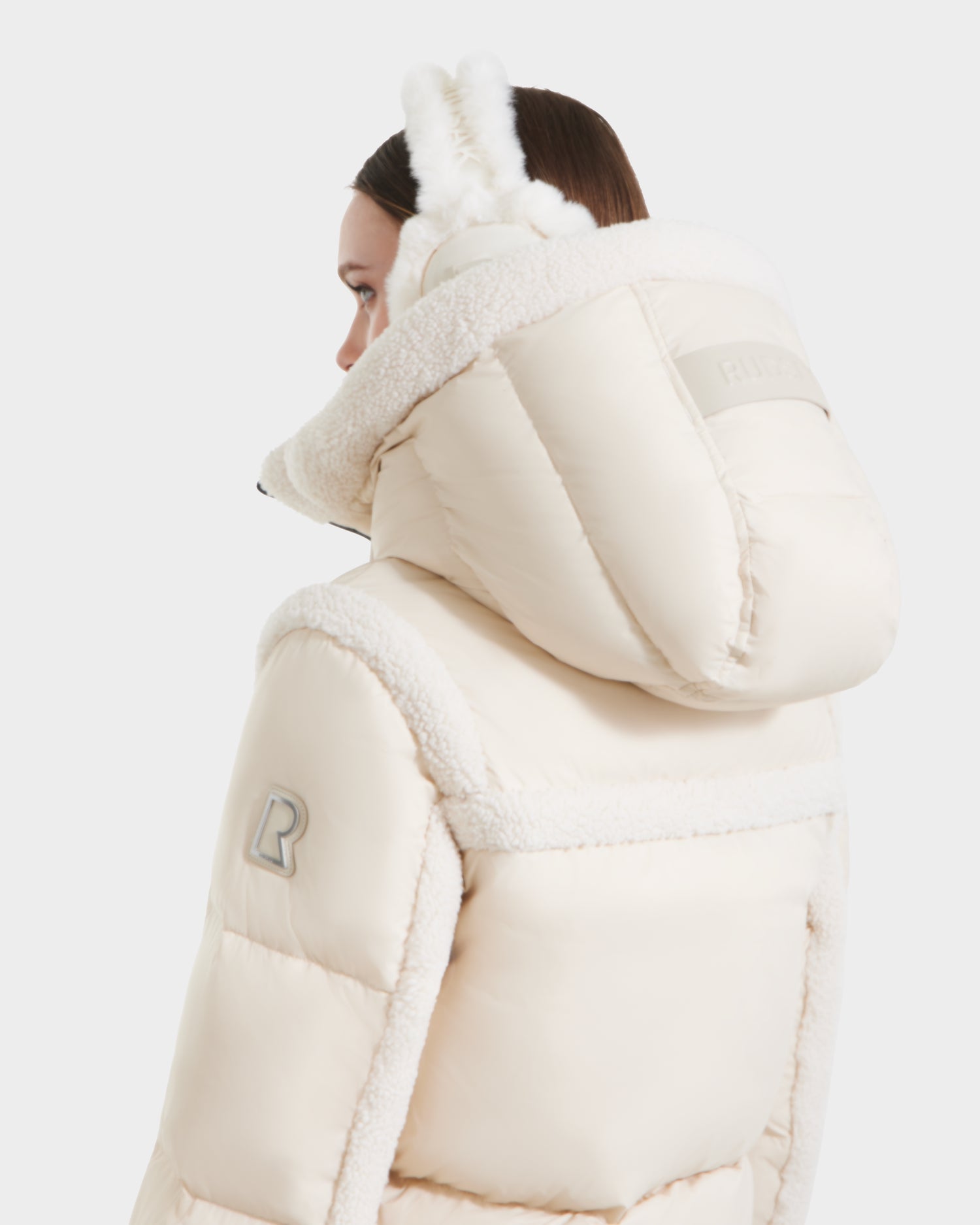 Wearing the Rudsak JULIANA - L124509 Vanilla women’s short down puffer with a large hood and shearling trim, a person holds fluffy earmuffs to their head, seen from the back and side against a white background.