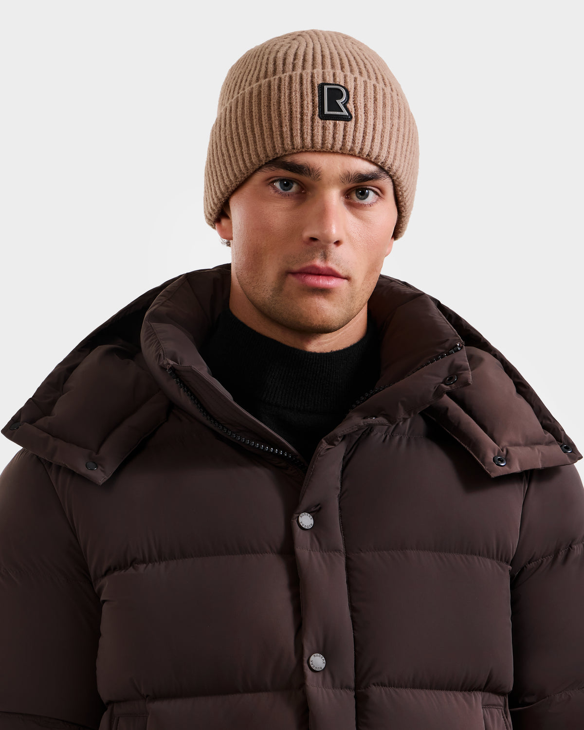 A person wears the Rudsak JULIO - U423502 Sand unisex knit beanie with a black patch and a dark brown puffer over a black turtleneck, standing against a light background for a classic winter essential look.