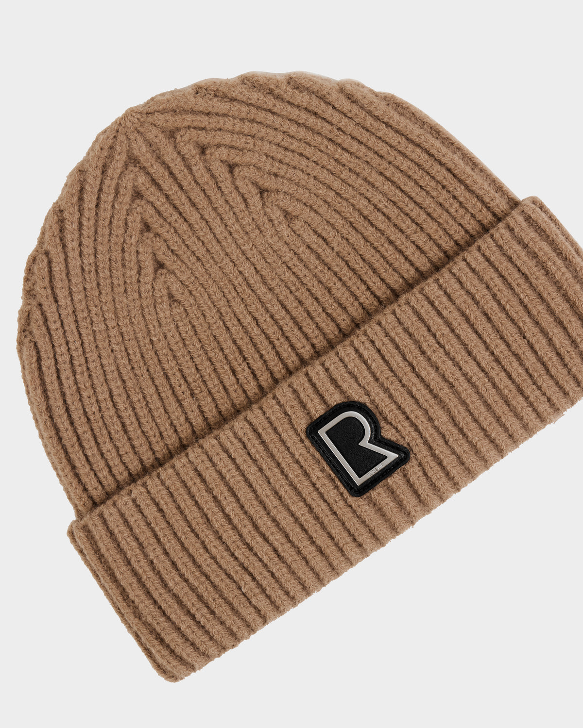 The Rudsak JULIO - U423502 Sand is a unisex knit beanie in brown with a folded brim and a black-and-white letter R patch on the front, shown on a white background—a must-have winter essential.