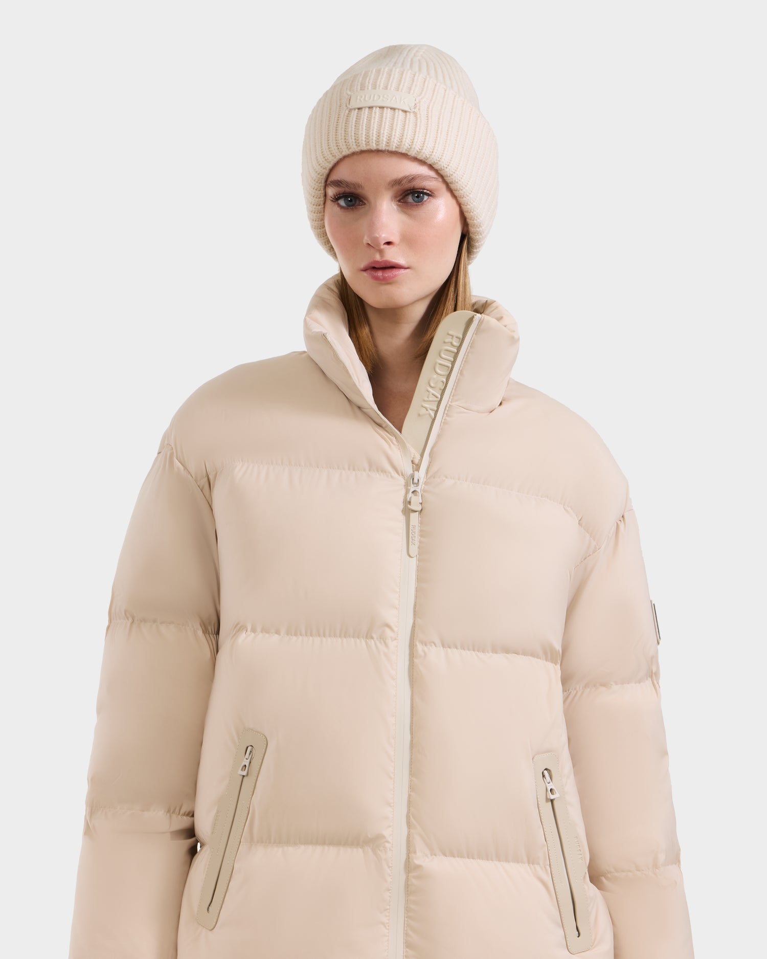 A woman wears the Rudsak KARLEE L - 8125682 Vanilla jacket with zippered pockets, vegan leather trims, and a high collar, paired with a matching knit beanie, posing against a plain white background.