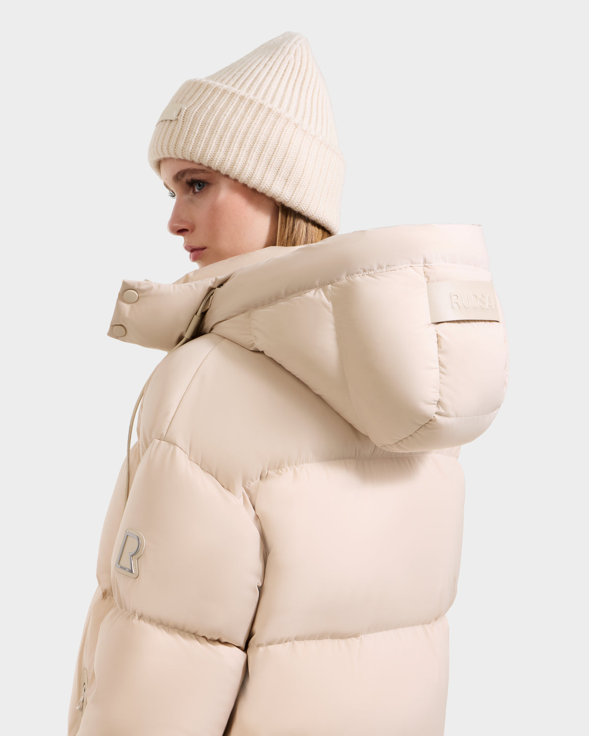 A woman in the Rudsak KARLEE L - 8125682 Vanilla winter jacket, featuring vegan leather trims and a large hood, is shown from the side wearing a cream-colored beanie, set against a plain light background.