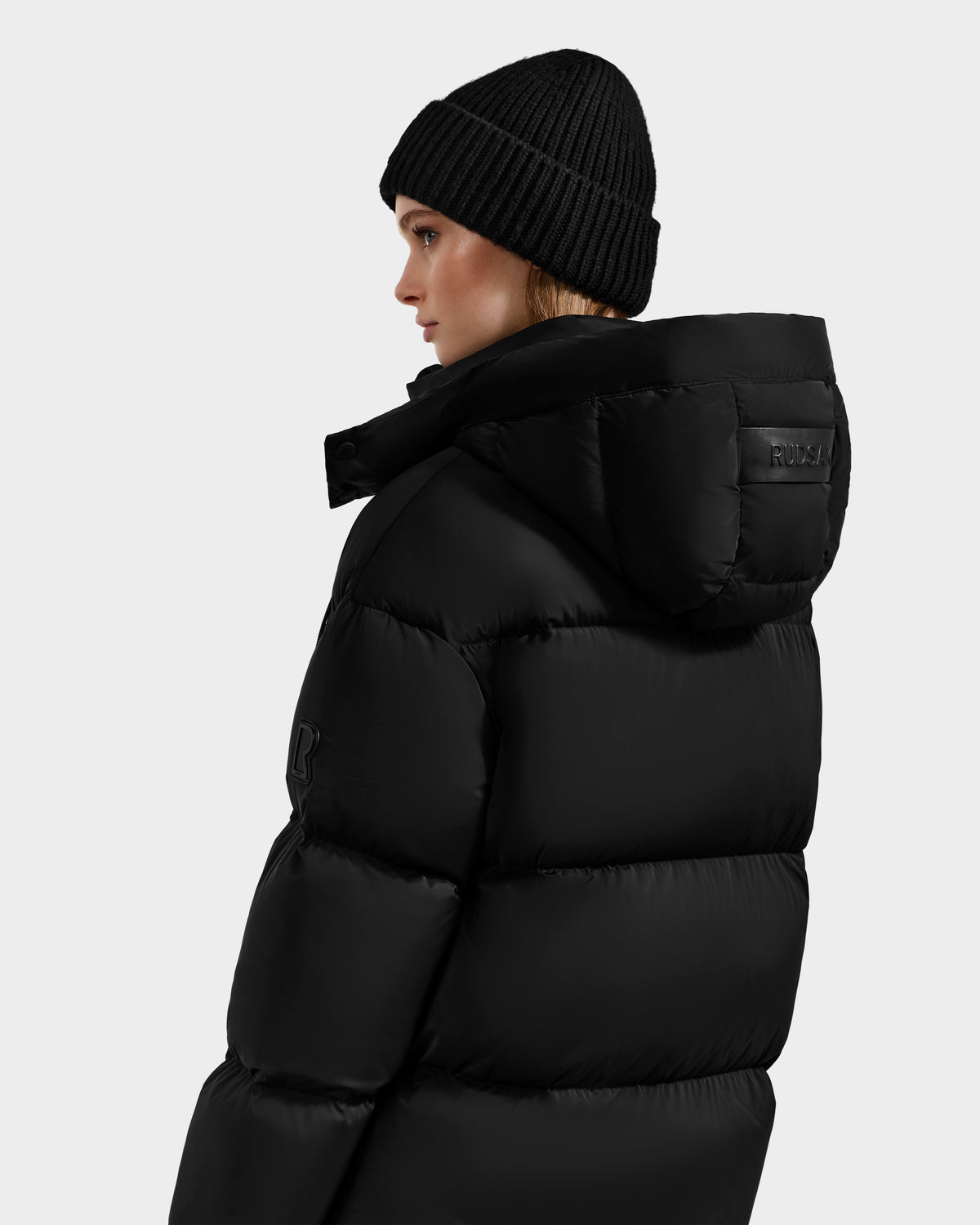 A woman models the Rudsak KARLEE XL - 8125683 Black beanie and women's maxi down puffer jacket, highlighting their style and warmth, shown from the side and back against a plain light grey background.