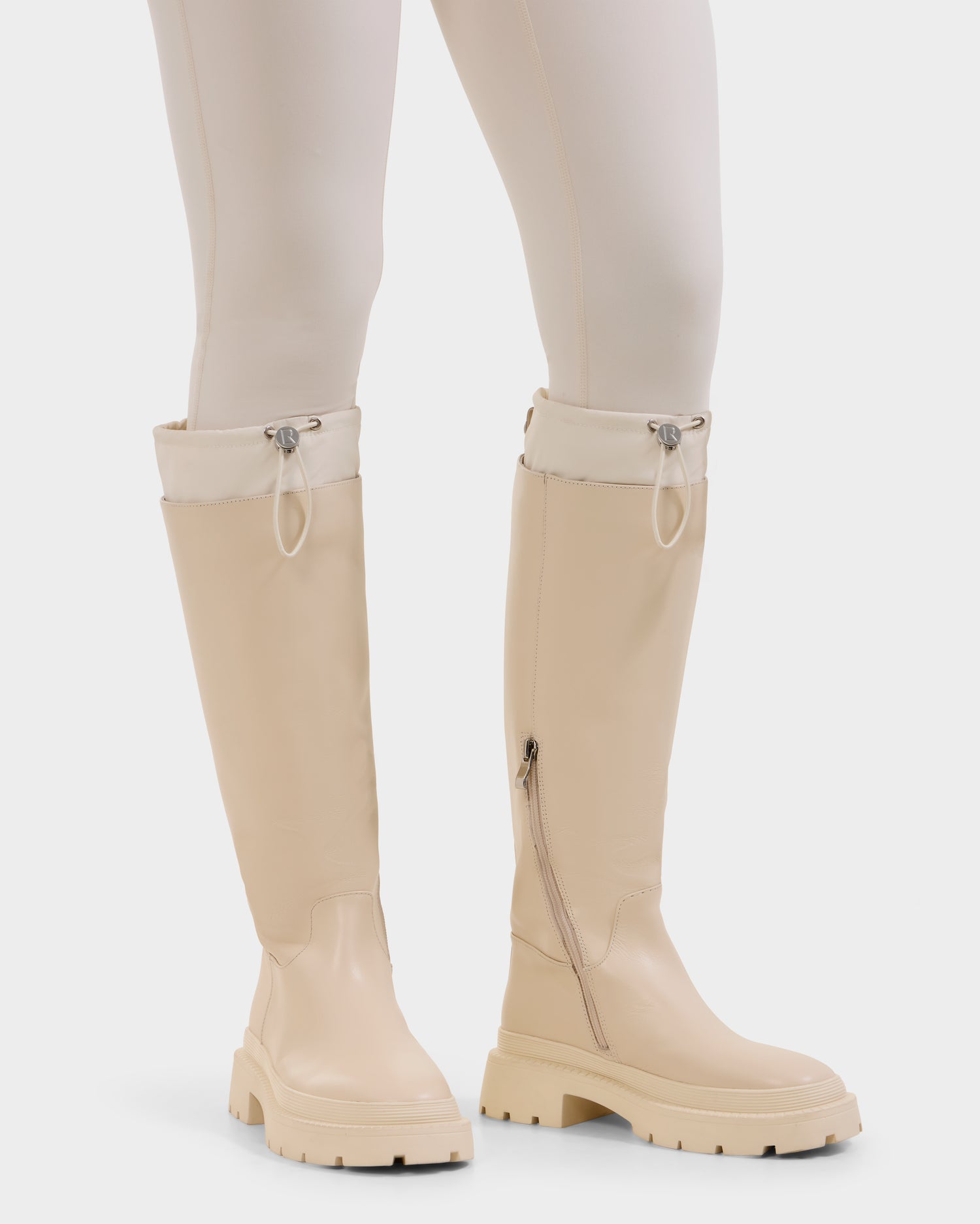 A person wears Rudsak's KAROL - 8225576 Vanilla women’s high leather boots with chunky rubber soles, side zippers, and a drawstring cuff, paired with matching cream fitted pants.