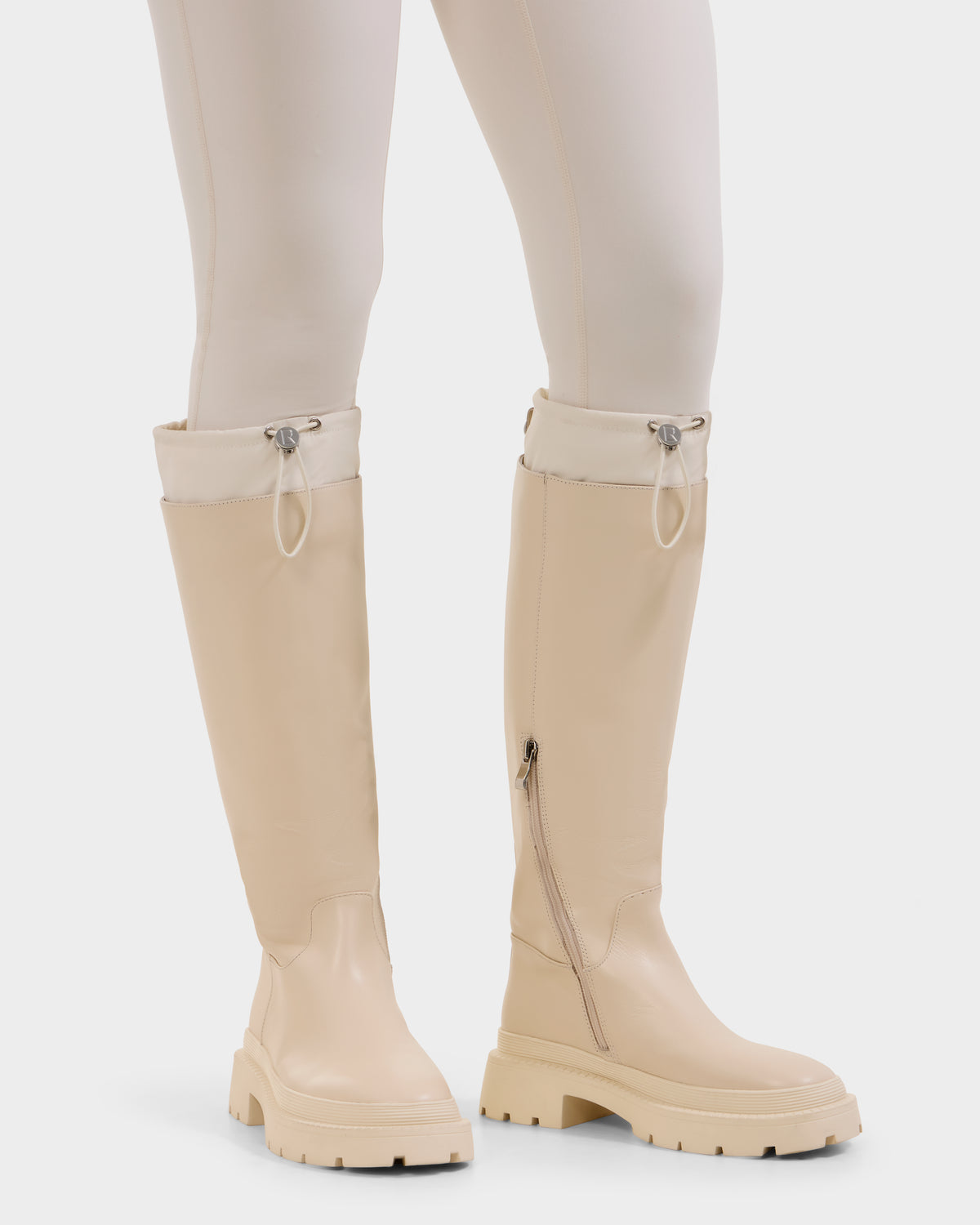 A person wears Rudsak's KAROL - 8225576 Vanilla women’s high leather boots with chunky rubber soles, side zippers, and a drawstring cuff, paired with matching cream fitted pants.