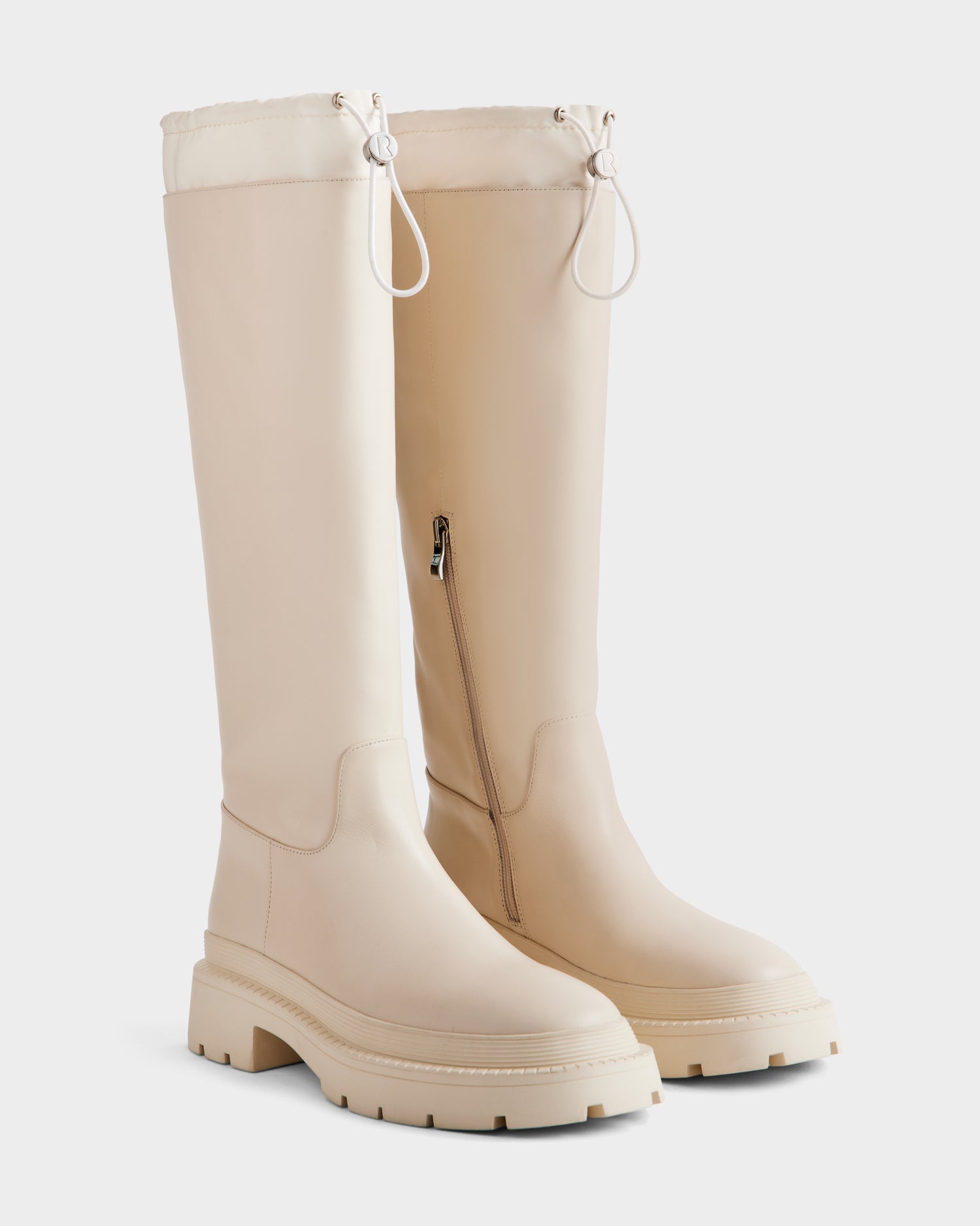 The KAROL - 8225576 Vanilla by Rudsak are knee-high, beige leather women’s boots with chunky rubber soles, side zippers, and adjustable cuffs. These modern boots are shown against a plain white background.