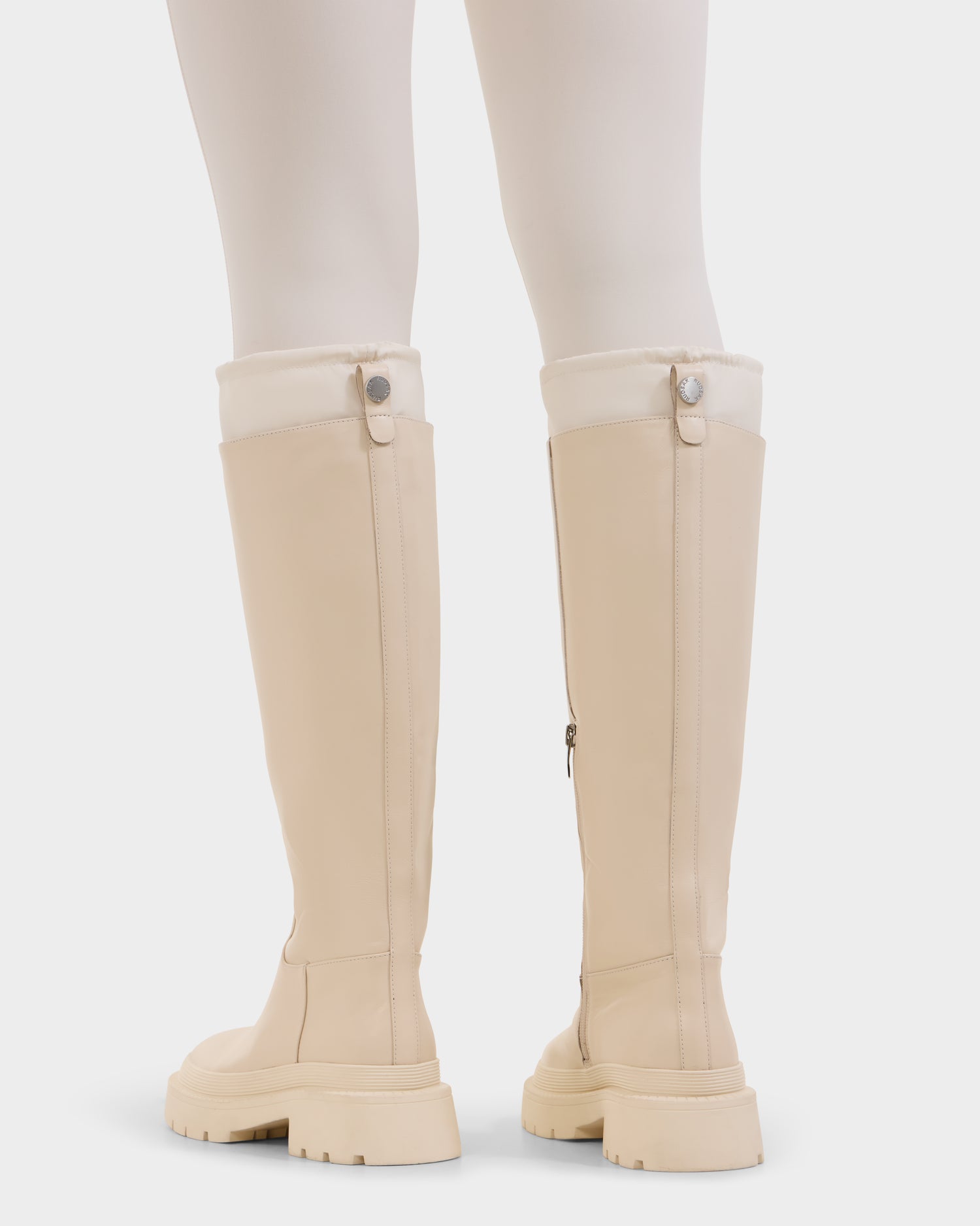 A person wearing Rudsak KAROL - 8225576 Vanilla cream-colored women’s high leather boots with chunky soles, white fleece trim, and an adjustable cuff, photographed from behind against a white background.