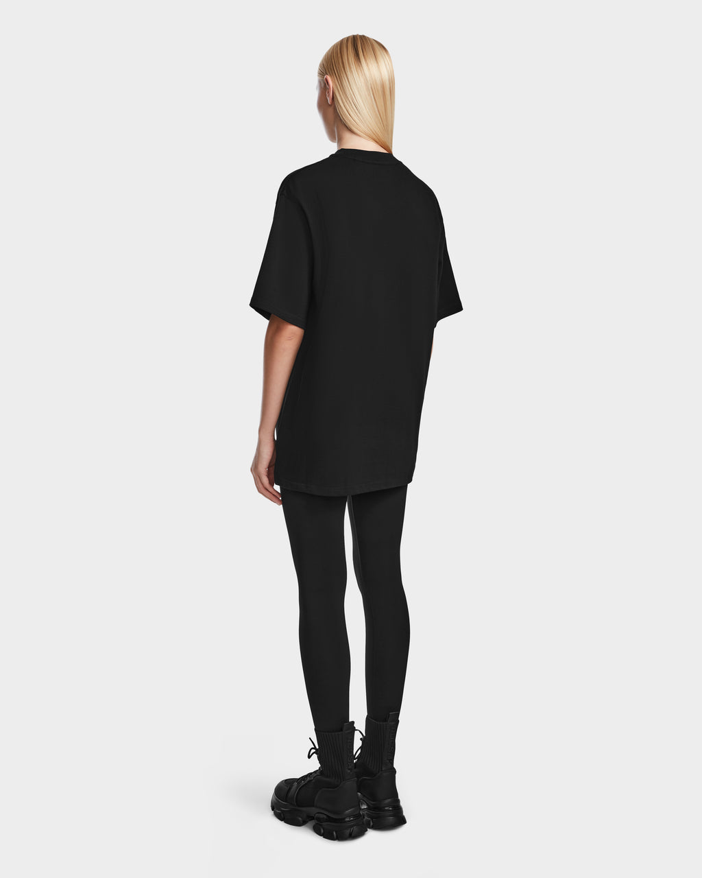 A person with long, straight blonde hair stands facing away from the camera, wearing the KARTER - 9524008 Black W oversized unisex T-shirt by Rudsak, paired with black leggings and sneakers against a light gray background.