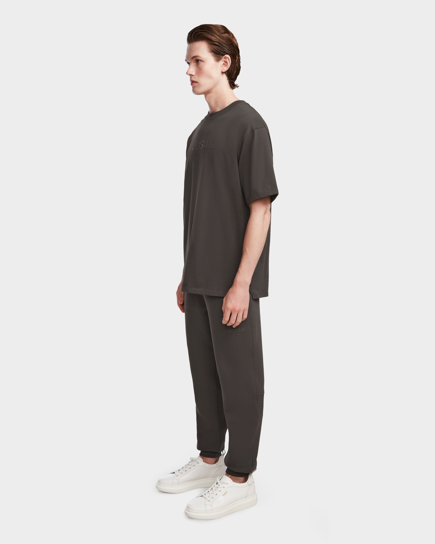 A man stands in profile against a white background, wearing the Rudsak KARTER - 9524008 Grey M relaxed fit unisex T-shirt, matching pants, and white sneakers, all crafted from premium cotton.