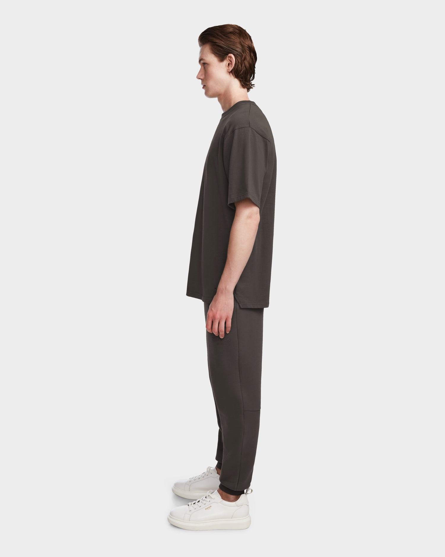 A man in profile models the Rudsak KARTER - 9524008 Grey M, a relaxed fit, oversized unisex premium cotton T-shirt with matching pants and white sneakers, set against a plain white background.