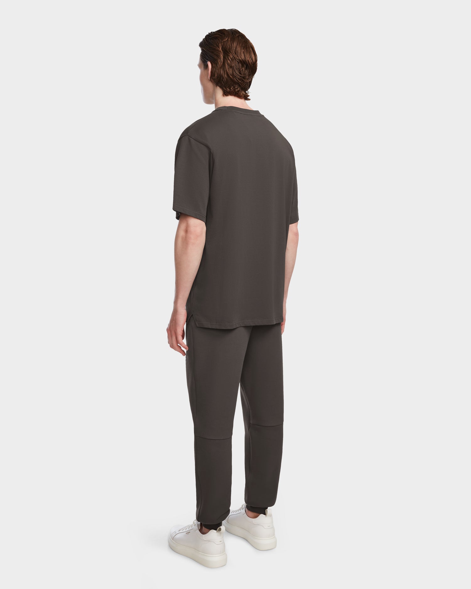A person with short brown hair faces away, wearing the Rudsak KARTER - 9524008 Grey M relaxed fit unisex T-shirt and matching pants with white sneakers, set against a plain white background.