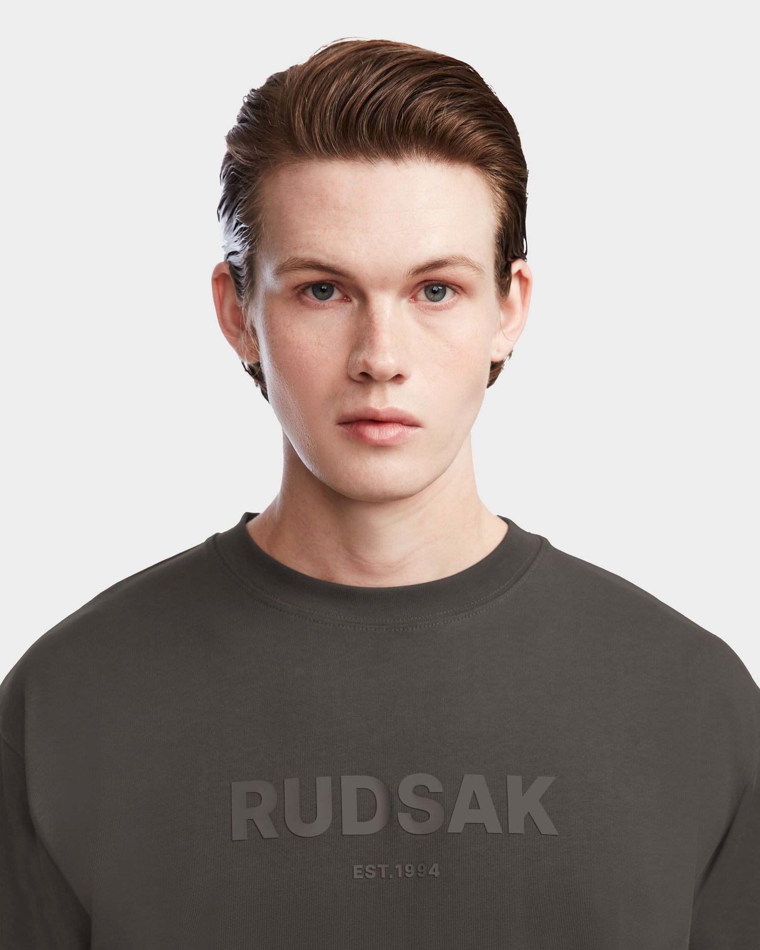Young man with fair skin and short brown hair wears the Rudsak KARTER - 9524008 Grey M relaxed fit sweatshirt, featuring RUDSAK EST. 1994 on the front, looking at the camera against a light background.