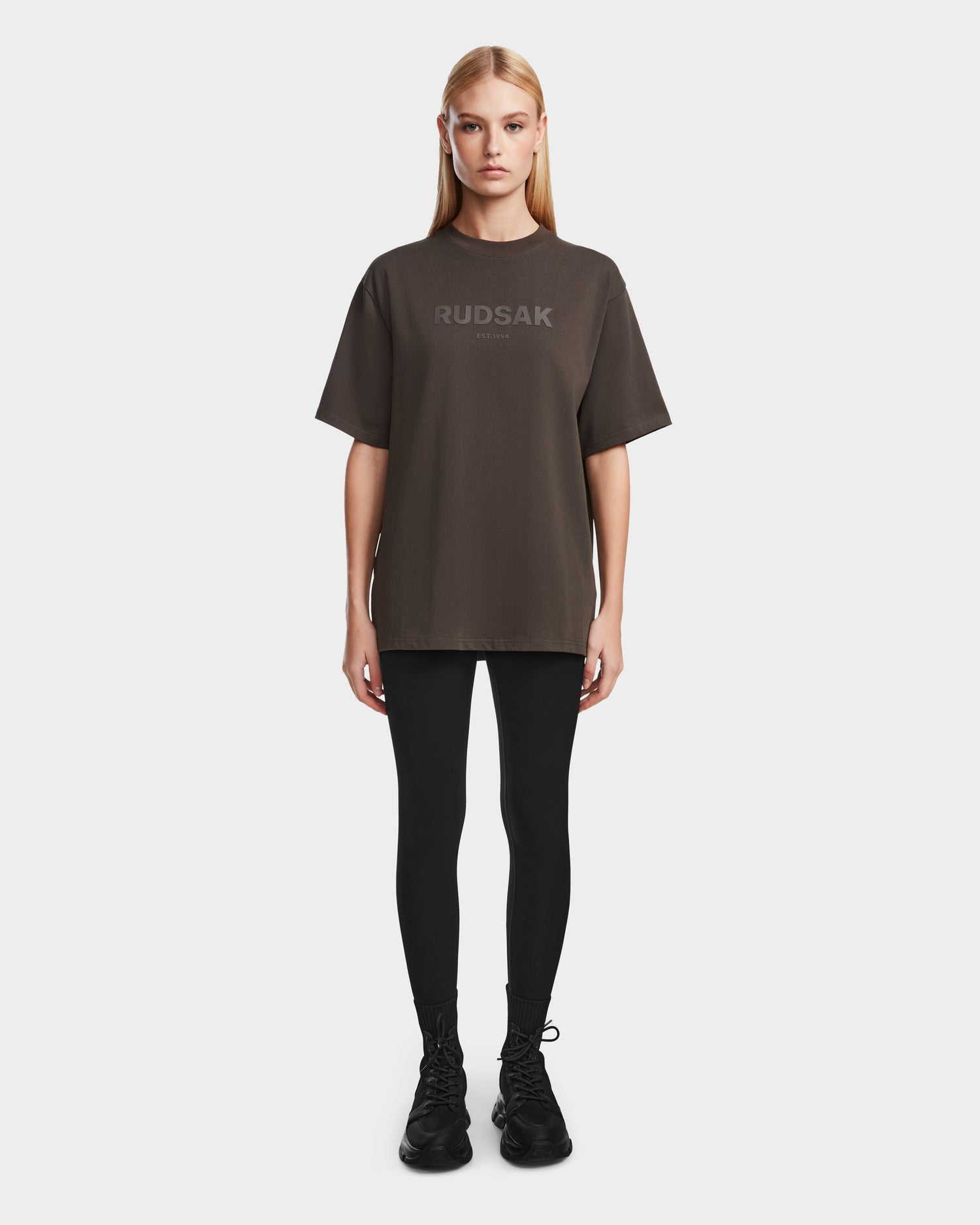 A woman stands facing forward, wearing a relaxed fit, oversized unisex Rudsak KARTER - 9524008 Grey W T-shirt, black leggings, and black shoes against a plain white background.