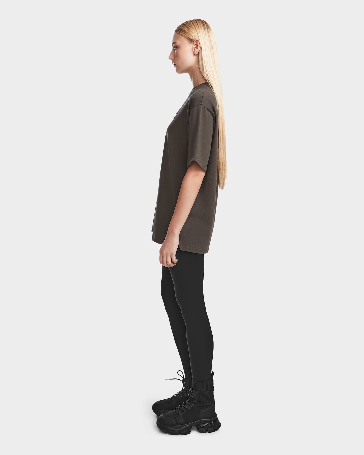 A woman with long blonde hair stands in profile against a plain background, wearing the Rudsak KARTER - 9524008 Grey W unisex oversized T-shirt in premium cotton, paired with black leggings and black lace-up boots.
