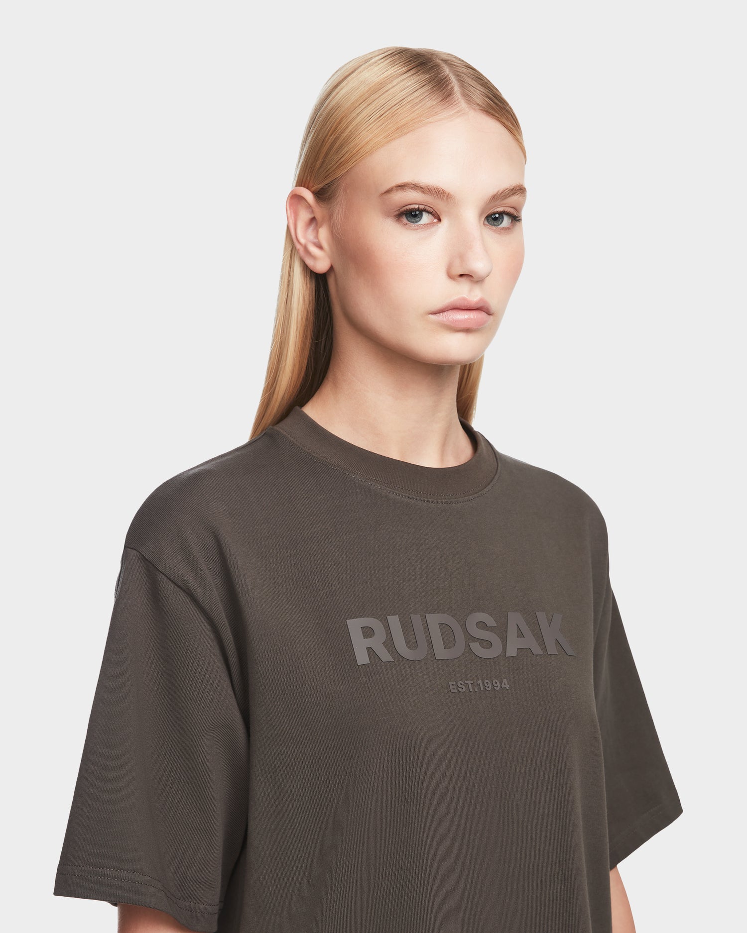 A woman with straight blonde hair wears the Rudsak KARTER - 9524008 Grey W oversized unisex T-shirt, made from premium cotton and featuring RUDSAK EST. 1994 on the front, standing against a plain light background.