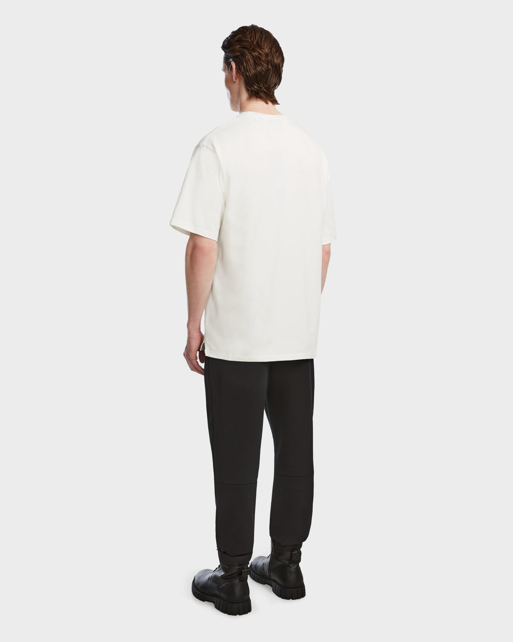 A person faces away, wearing the Rudsak KARTER - 9524008 Iceland M relaxed-fit unisex T-shirt in white, paired with black pants and chunky black boots against a plain light background.