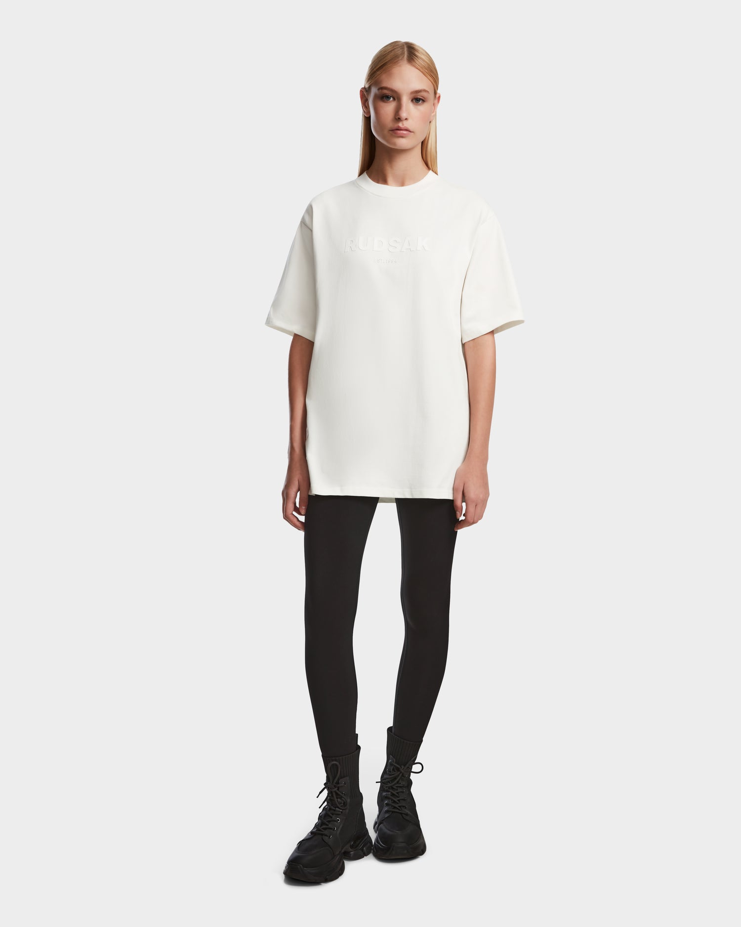 A woman with straight blonde hair wears the Rudsak KARTER - 9524008 Iceland W relaxed fit, oversized unisex premium cotton T-shirt, paired with black leggings and lace-up boots, standing against a plain white background.