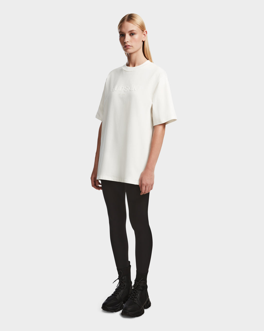 A person with long blonde hair stands against a plain background, wearing the Rudsak KARTER - 9524008 Iceland W oversized unisex T-shirt made from premium cotton, styled with black leggings and boots. The relaxed fit features subtle white chest text.