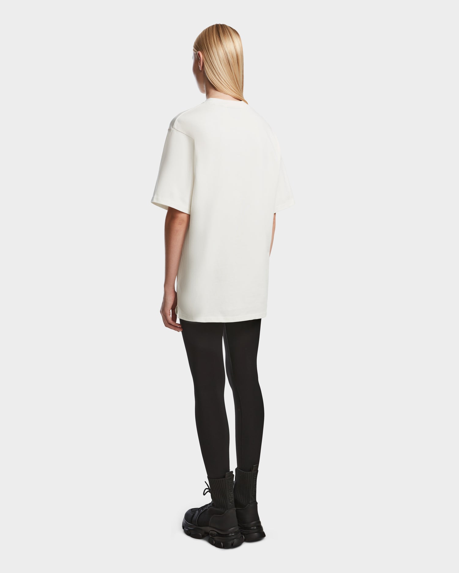 A person with straight blonde hair faces away, wearing the Rudsak KARTER - 9524008 Iceland W relaxed fit oversized unisex T-shirt in premium cotton, paired with black leggings and boots against a plain light background.