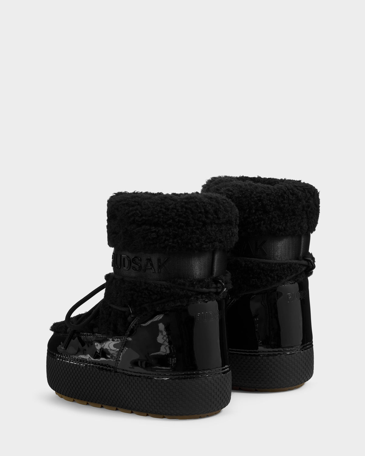 The Rudsak KATHY Y - 1224500 Black boots feature water-repellent shiny patent leather, textured cuffs, and lace-up details, shown from the back on a light gray background.
