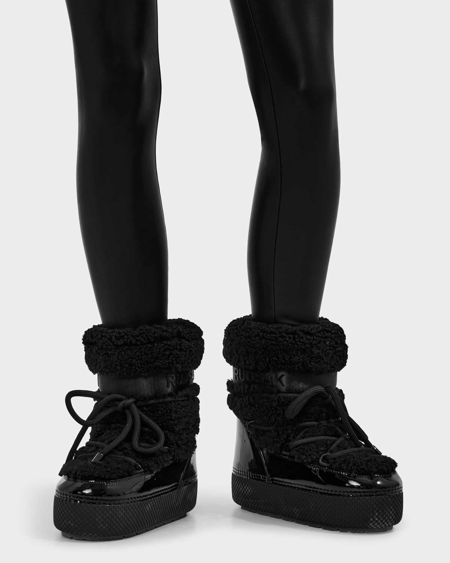 A person wearing Rudsak KATHY - 8223513 Black water-repellent boots with chunky soles and thick textured cuffs, paired with shiny black leggings, stands on a plain light background.