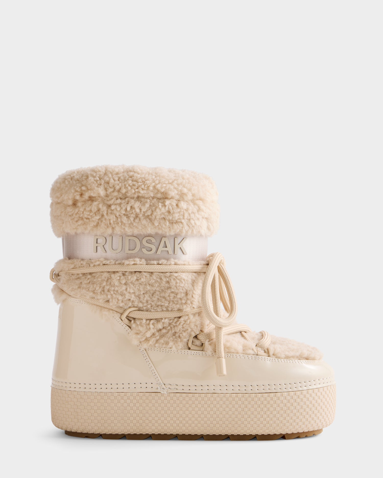 The KATHY - 8223513 Vanilla women’s winter boots by Rudsak feature a thick rubber sole, shiny patent finish, faux shearling upper, water-repellent design, laces, and RUDSAK branding on a fabric band near the top.