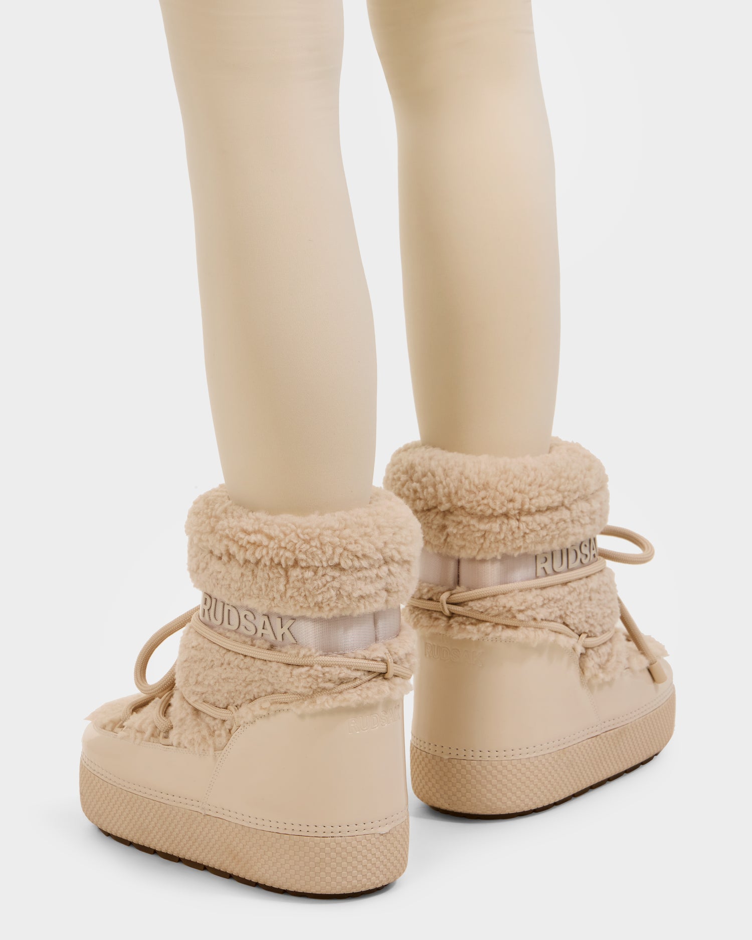 A person wearing cream tights and Rudsak KATHY - 8223513 Vanilla fur-lined beige winter boots with thick soles and laces, viewed from the back. The Rudsak branding appears on the upper part of the boots.