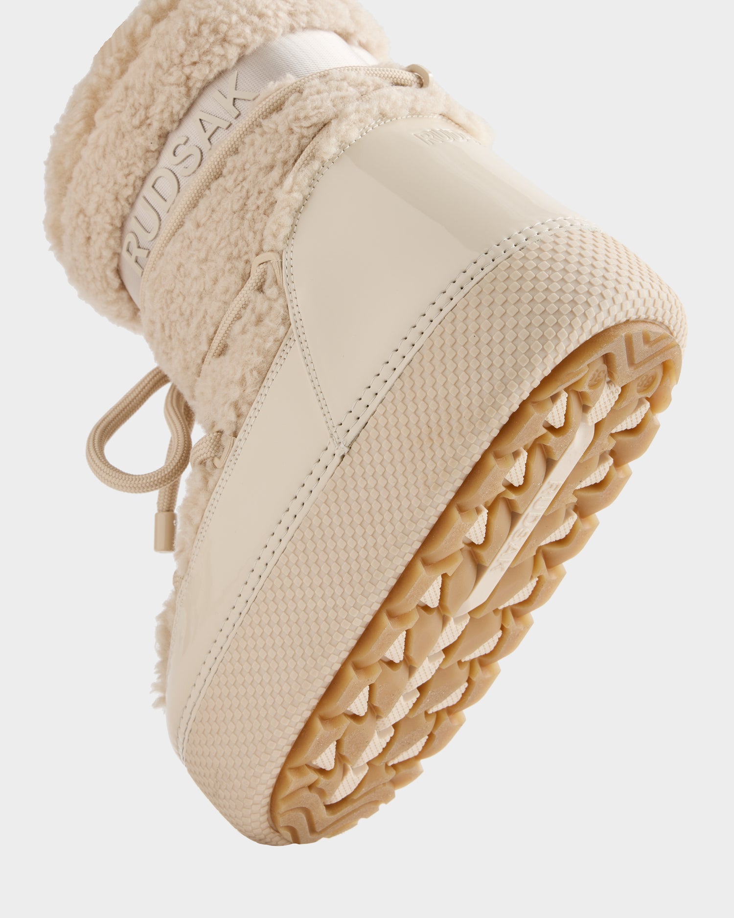 Close-up of the sole and lower part of the KATHY - 8223513 Vanilla women's winter boot by Rudsak, featuring a textured rubber outsole, patent leather, and fuzzy faux shearling upper. The RUDSAK logo is displayed on the side.