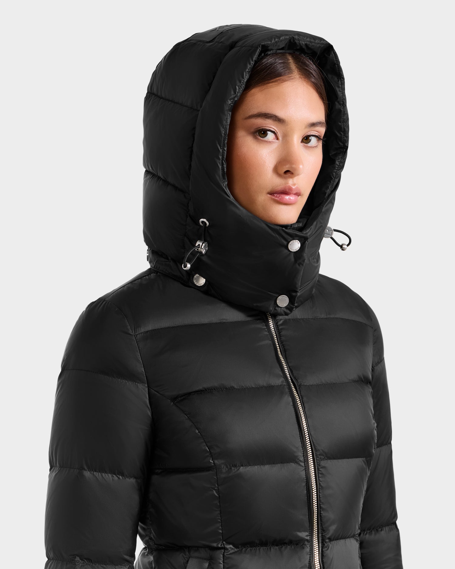 A woman in the Rudsak KATIA - L125564 Black puffer jacket with hood up, faces slightly sideways. The weather-resistant everyday jacket features a front zipper and buttoned hood. Background is light gray.