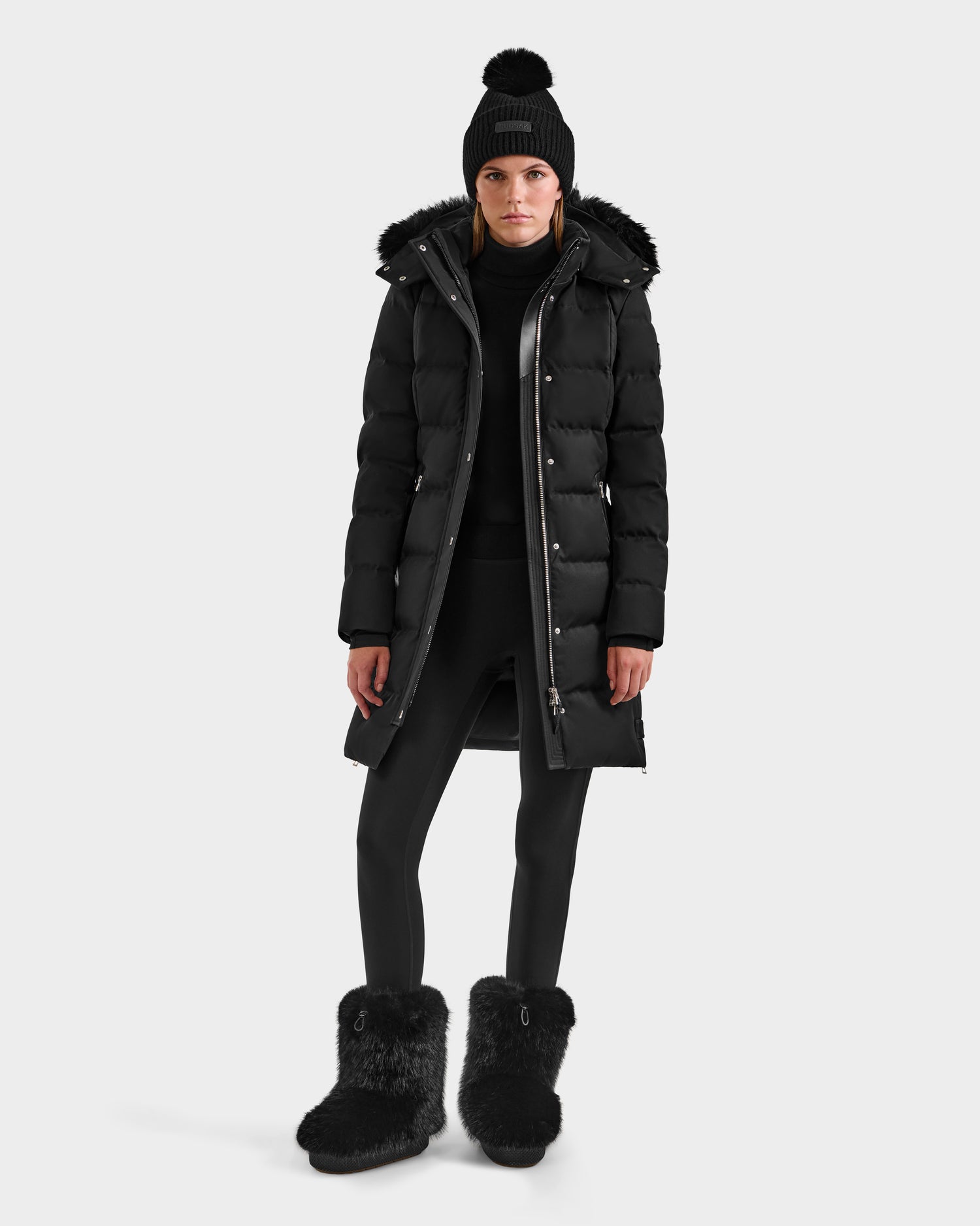 A person wears the Rudsak KEHLANI H - 8125643 Black women's down parka with fur-lined hood, paired with black leggings, a knit pom-pom beanie, and black fur boots, standing against a plain white background.
