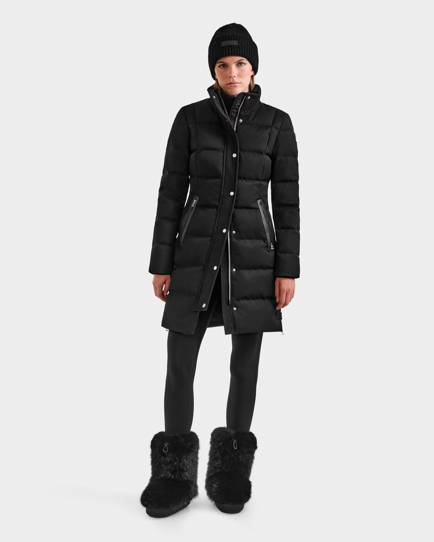A woman in the Rudsak KEHLANI H - 8125643 Black RDS-certified down coat, black leggings, fur boots, and a black beanie stands facing forward against a white background.