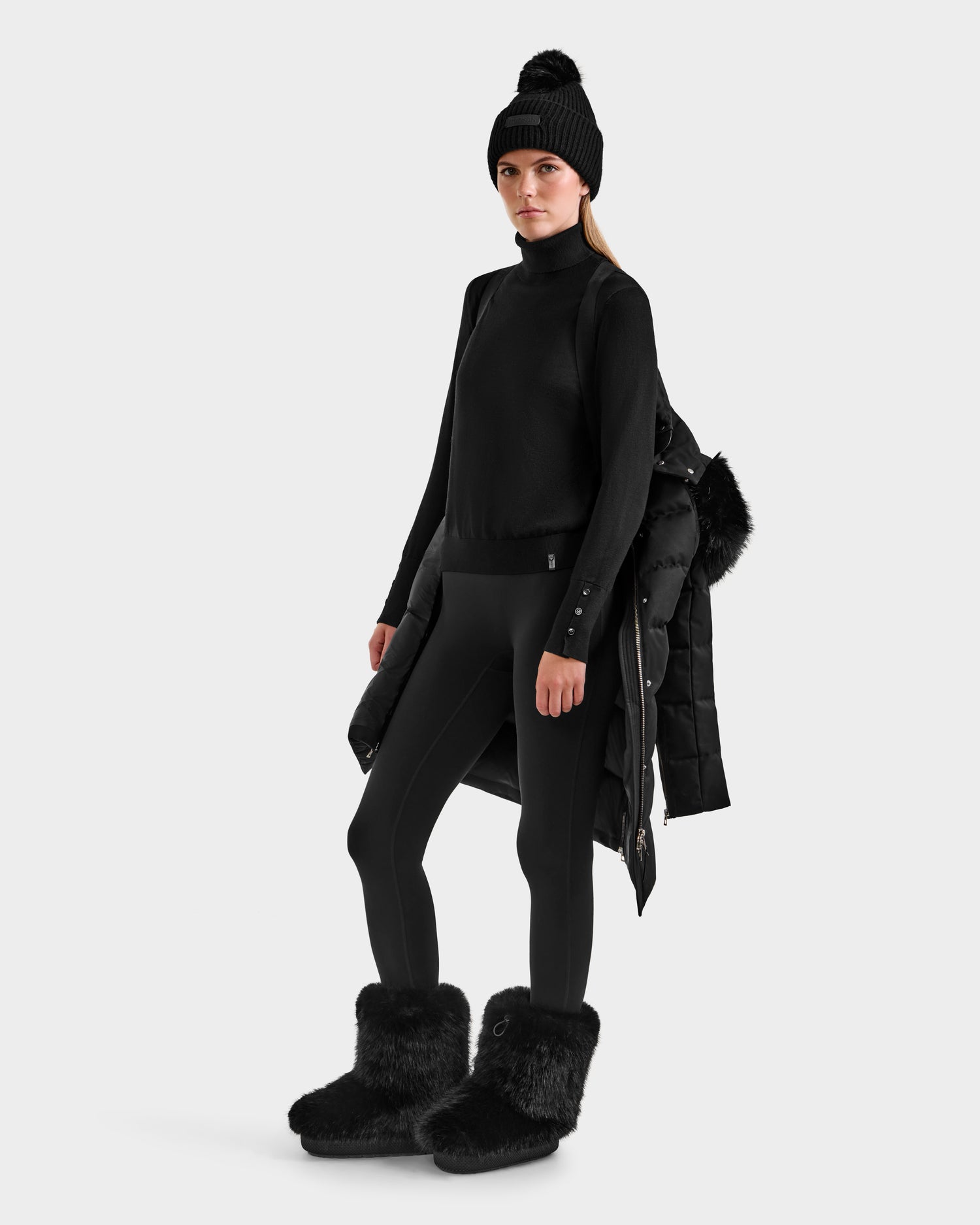 A woman in a black beanie, turtleneck, leggings, and furry boots stands with a Rudsak KEHLANI H - 8125643 Black women’s down parka, made from RDS-certified down, draped over her arm against a plain light background.