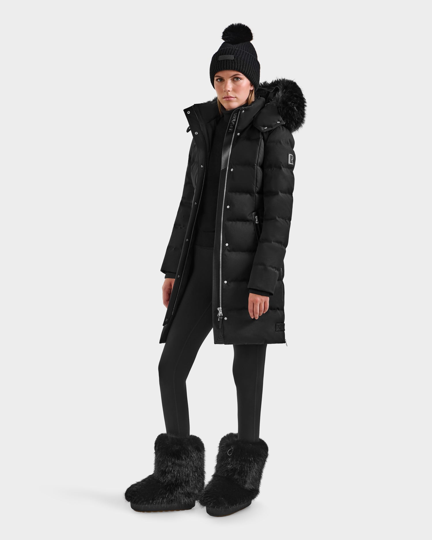 A person stands against a plain background wearing the Rudsak KEHLANI H - 8125643 black women's down parka with a fur-trimmed hood, paired with black leggings, fuzzy black boots, and a black knit pom-pom beanie.