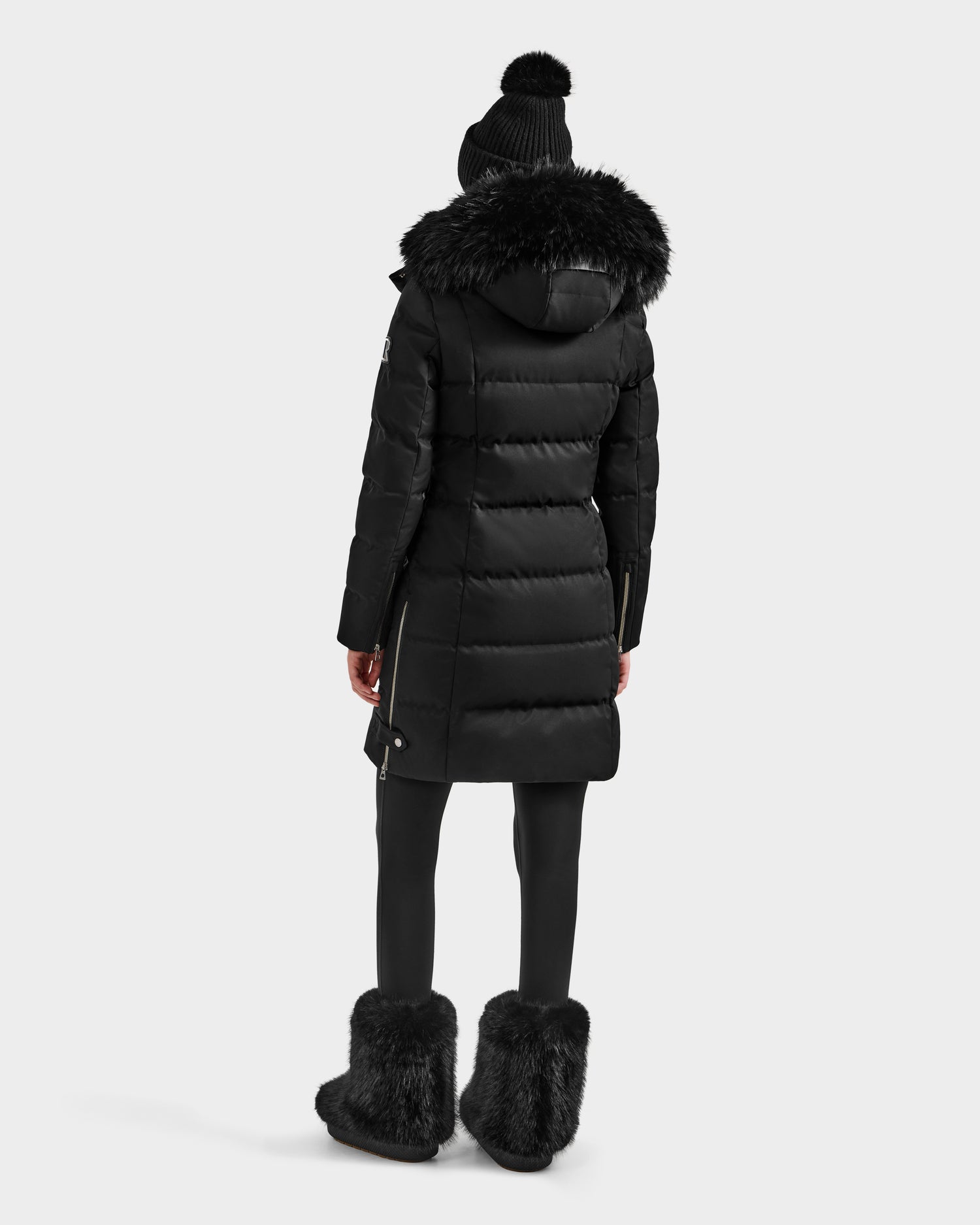 A person wears the Rudsak KEHLANI H - 8125643 Black women’s down parka with a fur-trimmed hood, paired with black leggings, fur boots, and a knit hat, standing against a plain light background.