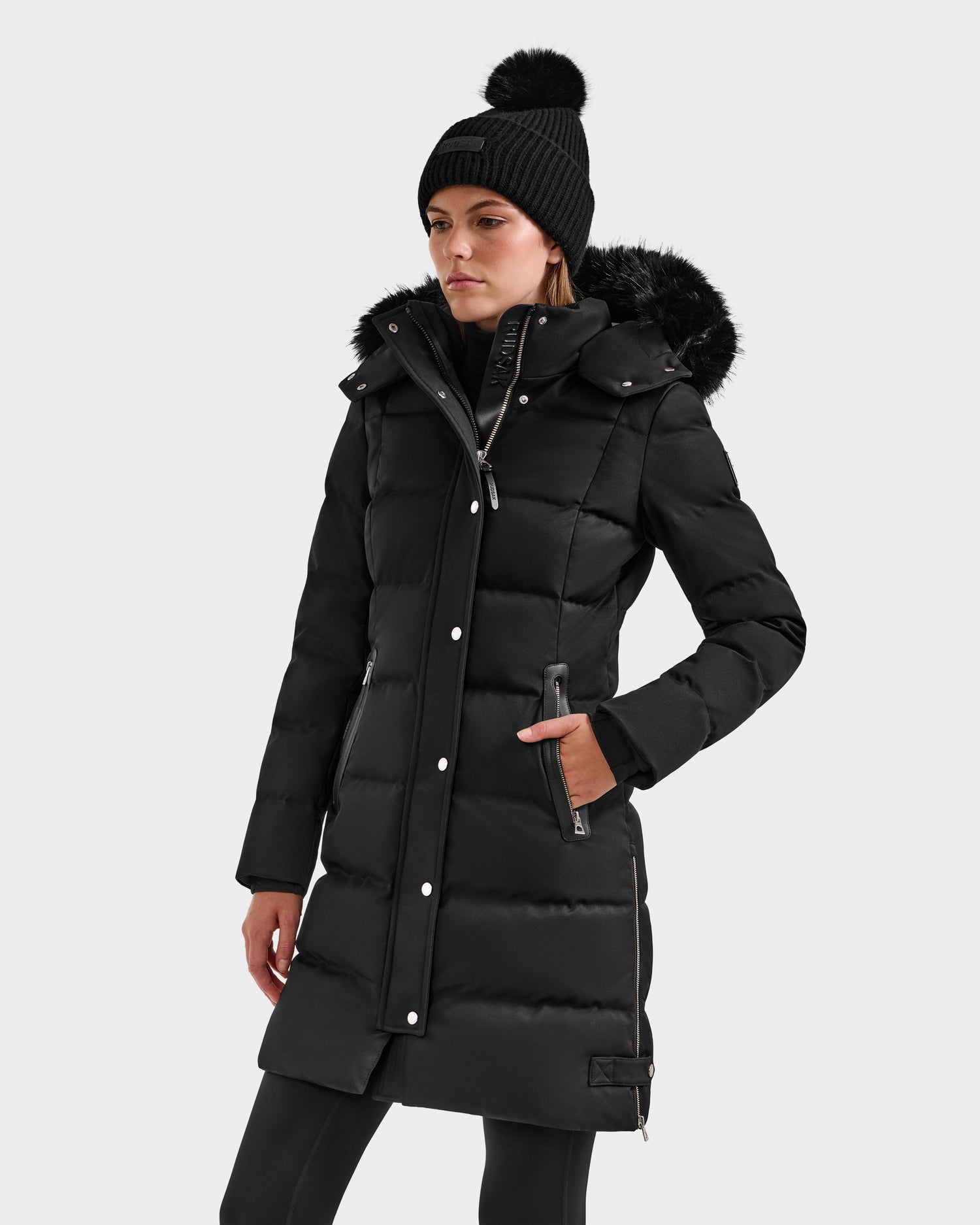A woman stands against a plain light background wearing the Rudsak KEHLANI H - 8125643 Black down parka with fur-lined hood and silver buttons, paired with a black pom-pom beanie and black pants.