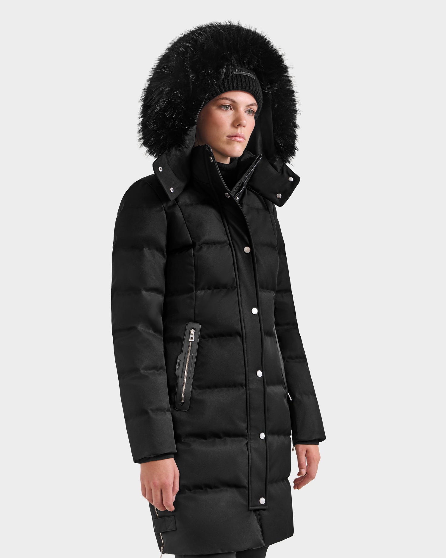 A woman wears the Rudsak KEHLANI H - 8125643 black winter coat with a large faux fur–trimmed hood and silver buttons, standing against a plain light gray background.