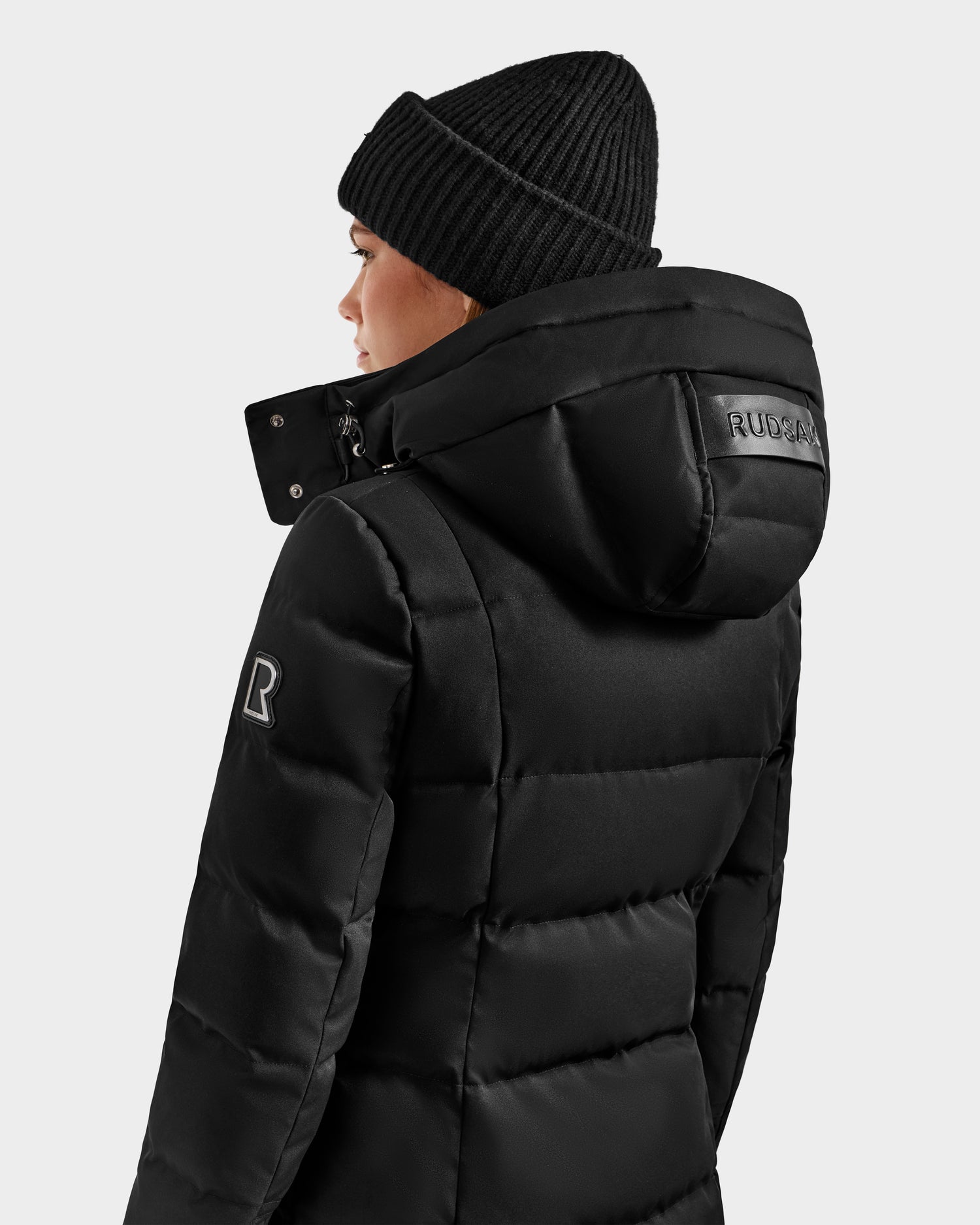 A person wearing the Rudsak KEHLANI H - 8125643 Black women’s down parka with a hood and black knit beanie, seen from the back and side. The winter coat features an R patch on the sleeve, "RUDSAK" on the hood, and is made with RDS-certified down.