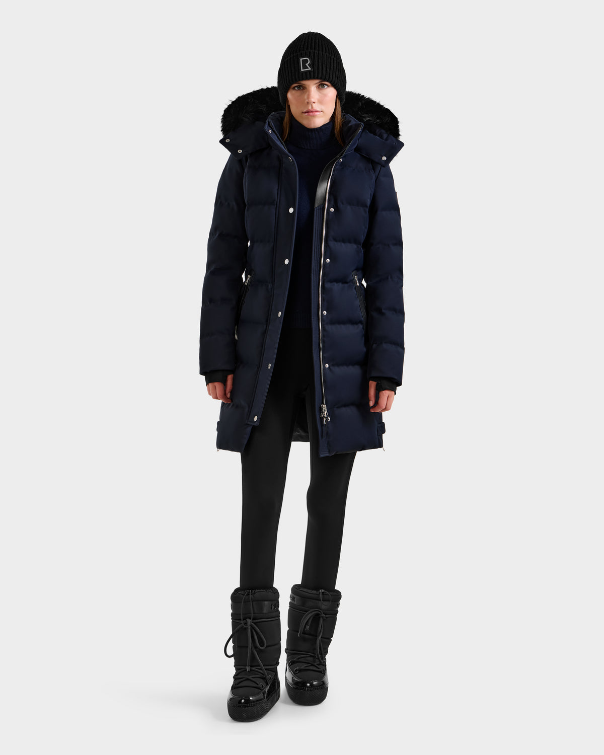 A woman faces forward in a Rudsak KEHLANI H - 8125643 Navy down parka with fur-trimmed hood, paired with black leggings, winter boots, and a knit beanie against a plain white background.