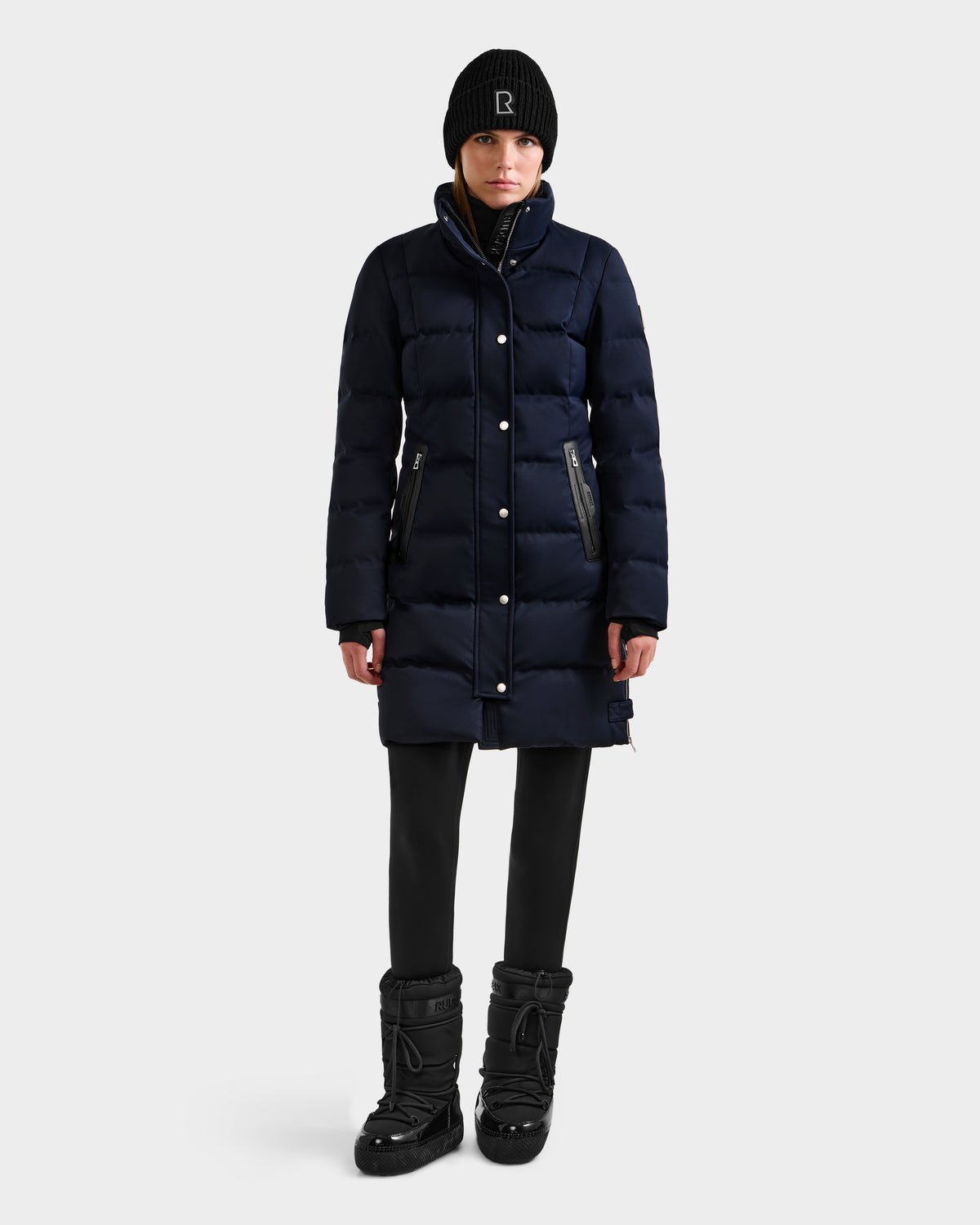 A person wearing the Rudsak KEHLANI H - 8125643 Navy women's down parka, black leggings, winter boots, and a black beanie stands facing forward against a plain white background.