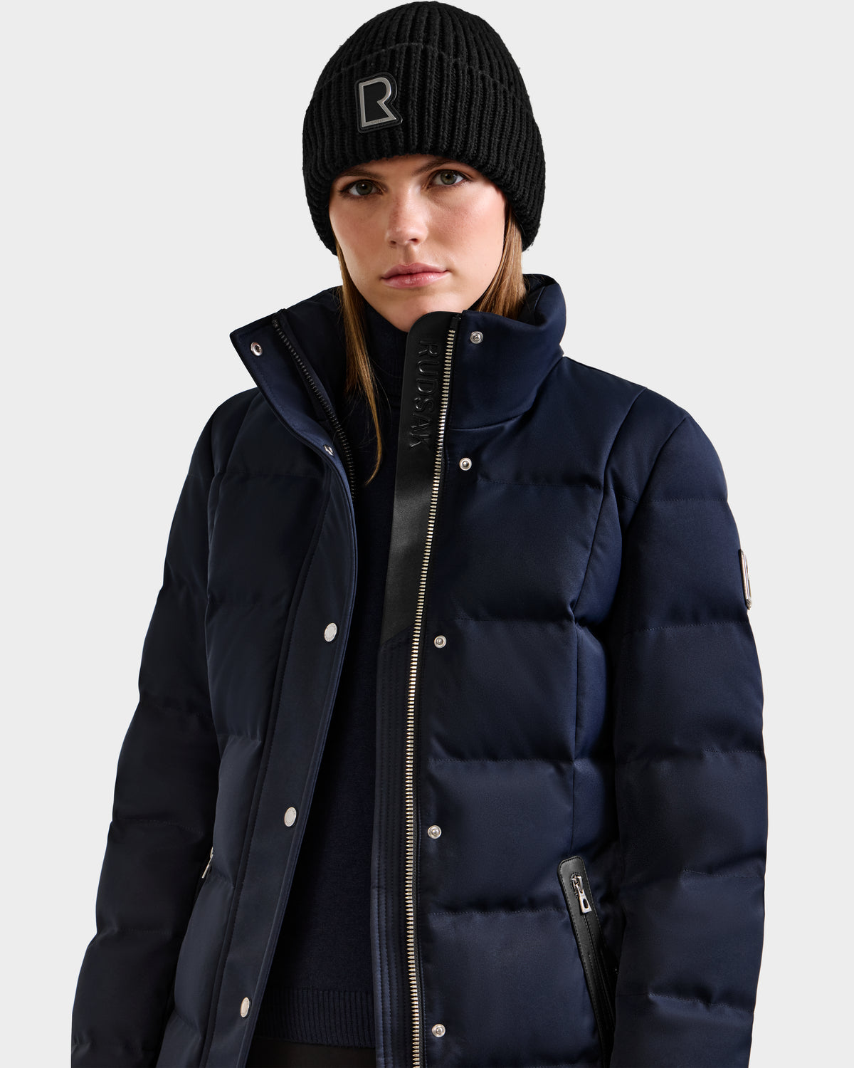 A person wearing the Rudsak KEHLANI H - 8125643 Navy women’s down parka with a high collar and a black knit beanie with a large R logo, standing against a light gray background.