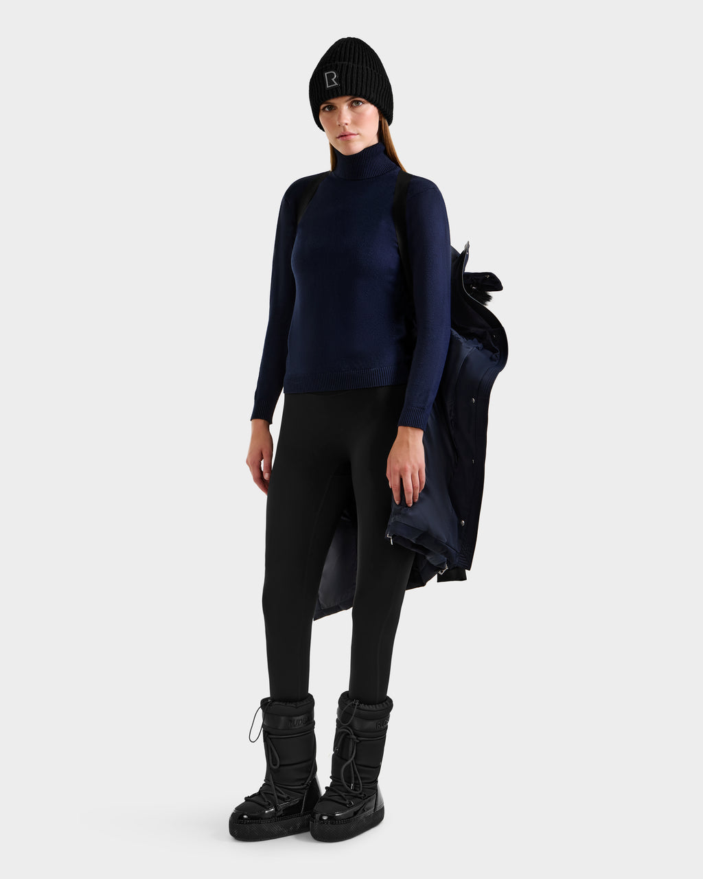A woman in a black beanie, navy turtleneck, black leggings, and snow boots stands holding the Rudsak KEHLANI H - 8125643 Navy RDS-certified down winter coat draped over her arm against a plain white background.