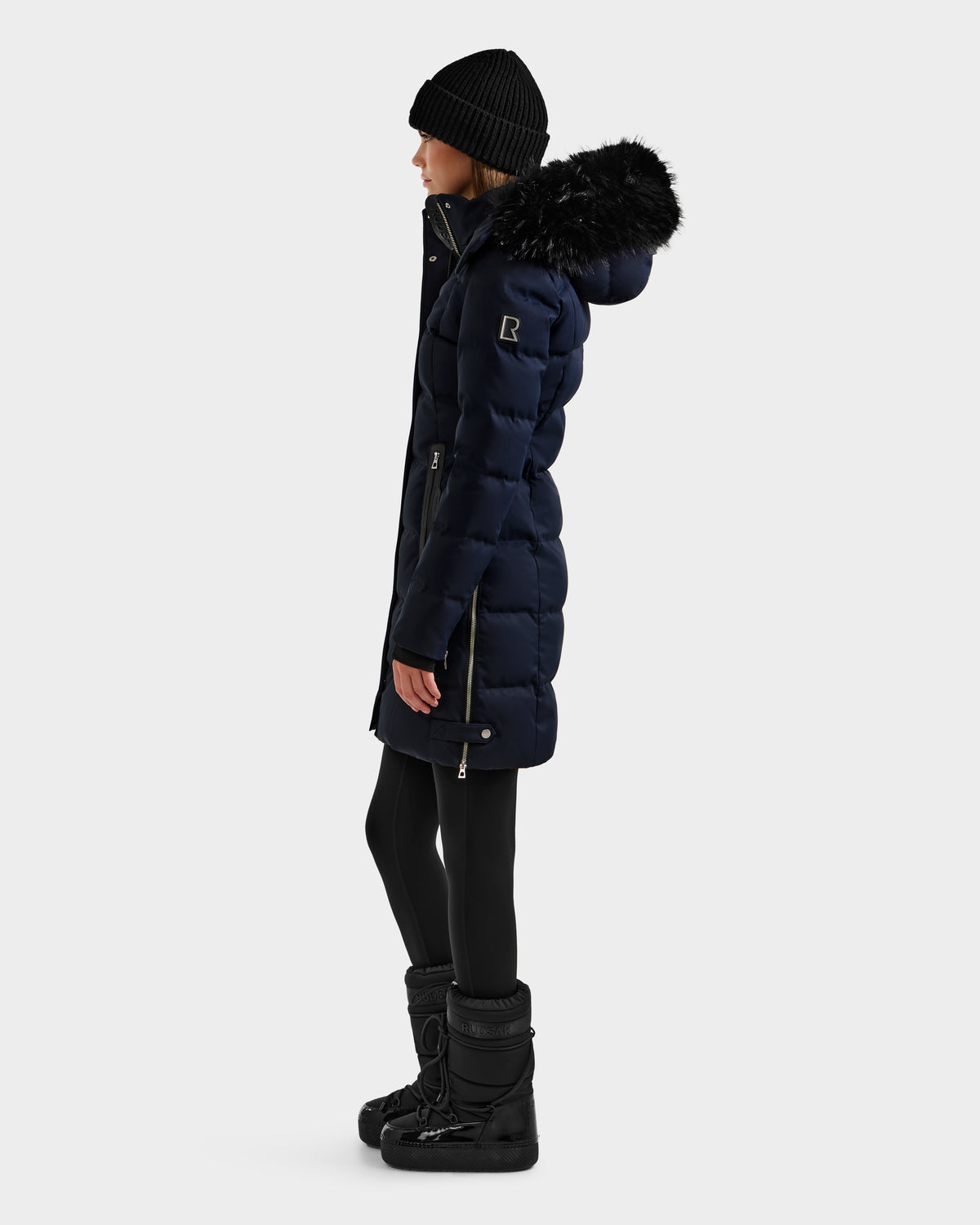 A person in the Rudsak KEHLANI H - 8125643 Navy women’s down parka with a fur-lined hood, black beanie, leggings, and winter boots stands in profile against a white background. The coat features RDS-certified down for extra warmth.