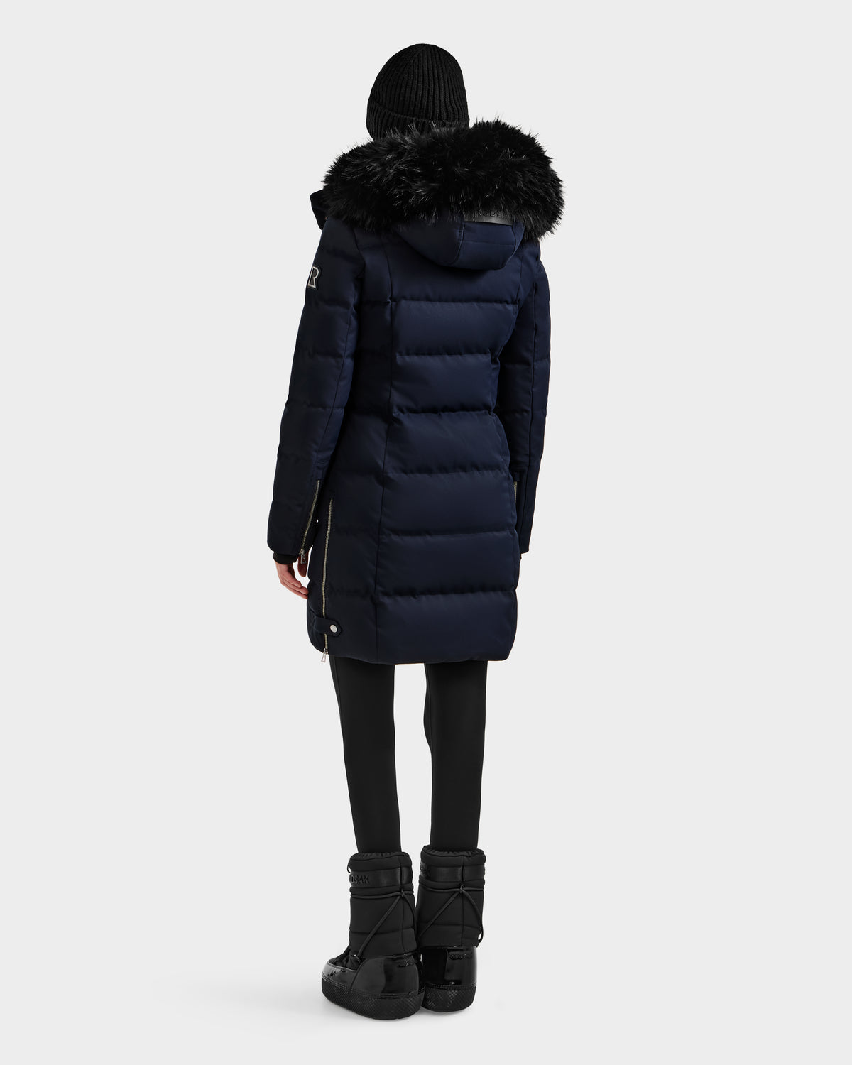 A person stands facing away, wearing the Rudsak KEHLANI H - 8125643 Navy women’s down parka with a black fur-lined hood, paired with black leggings, winter boots, and a knit beanie against a plain light background.