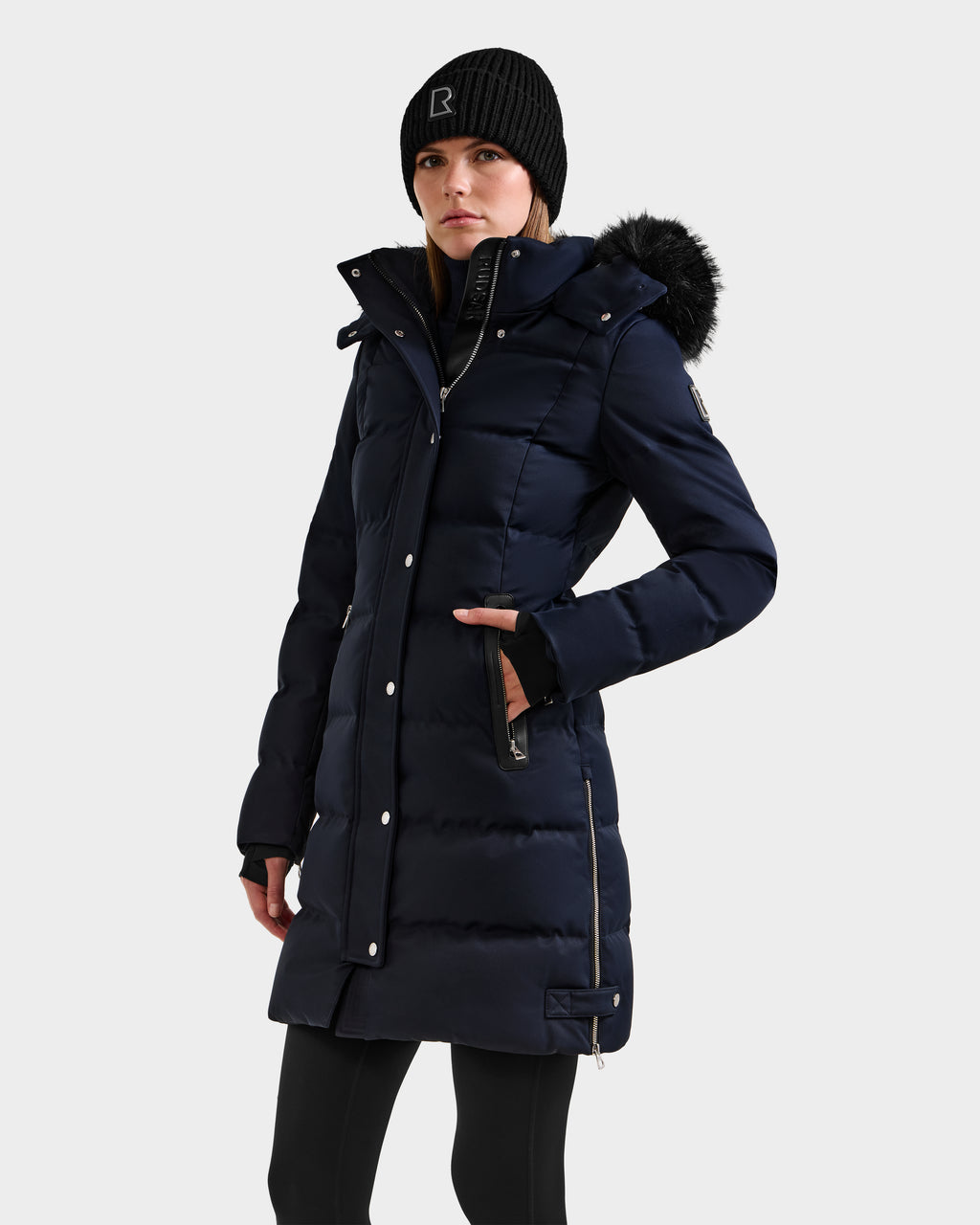 A woman wears the Rudsak KEHLANI H - 8125643 Navy women’s down parka with a fur-lined hood, paired with a black knit beanie and black leggings, standing against a plain light background.