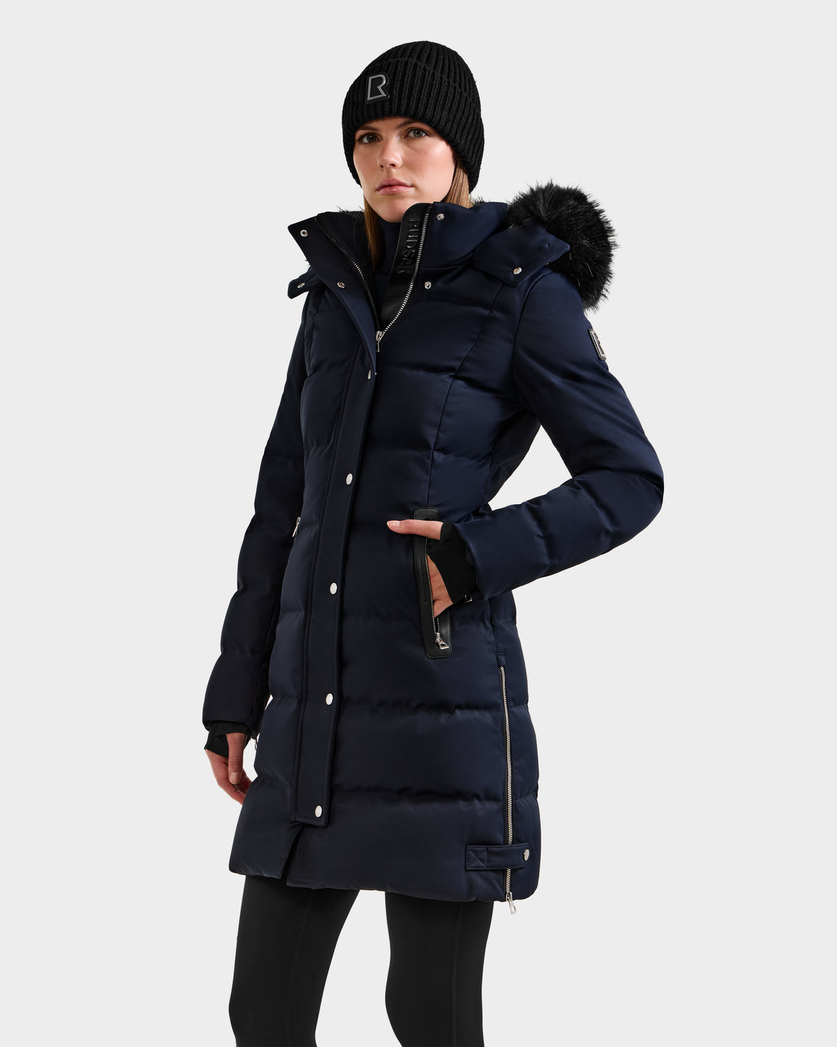 A woman wears the Rudsak KEHLANI H - 8125643 Navy women’s down parka with a fur-lined hood, paired with a black knit beanie and black leggings, standing against a plain light background.