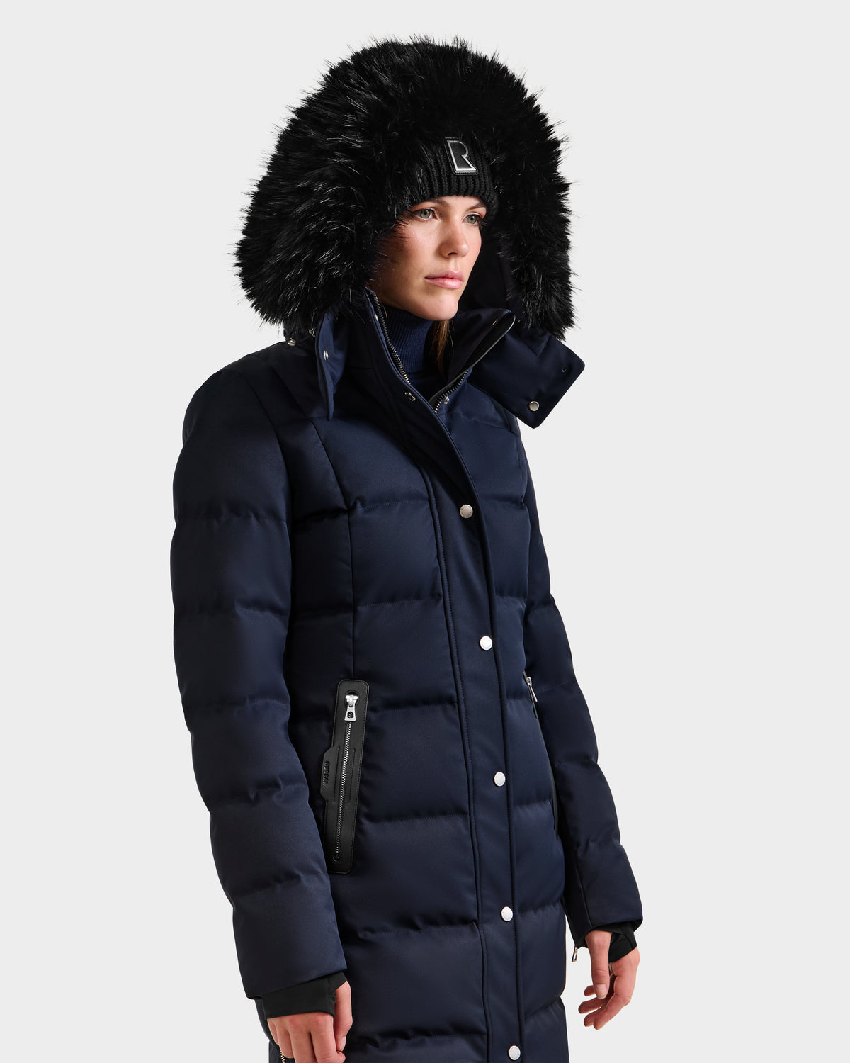 A person in the Rudsak KEHLANI H - 8125643 Navy long down coat with a faux fur-trimmed hood and black beanie stands against a plain light background.