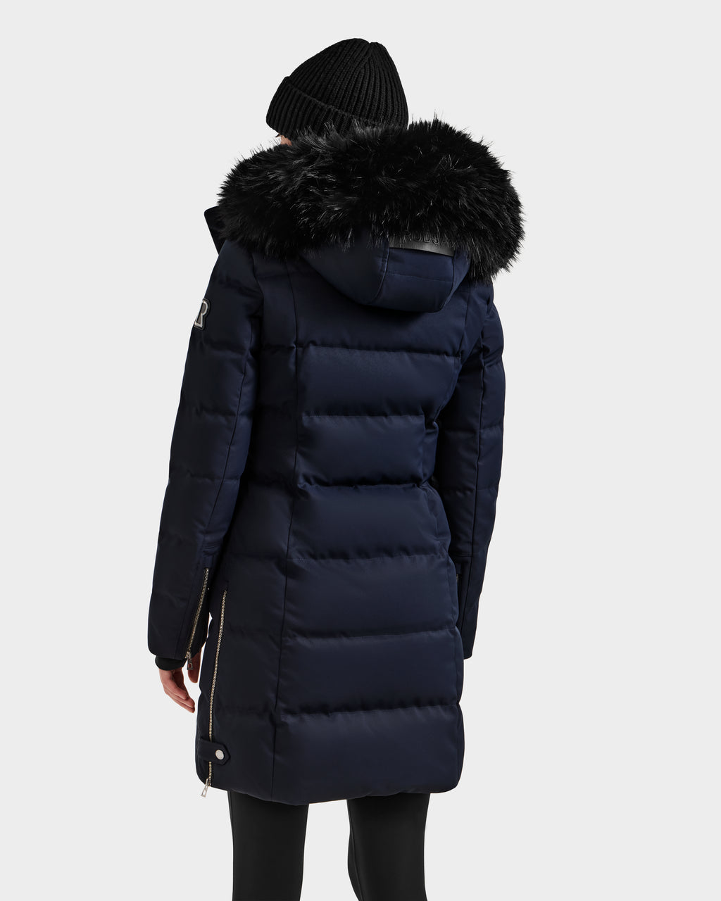 A person wearing a black knit beanie and the Rudsak KEHLANI H - 8125643 Navy women's down parka with a large black faux fur-trimmed hood is seen from the back against a plain light background.