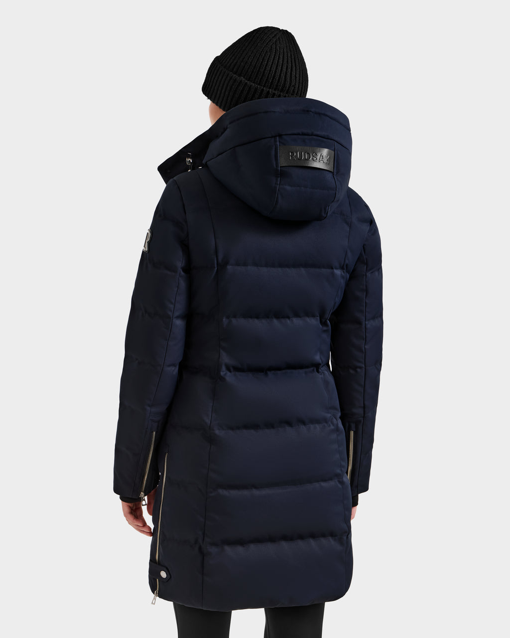 A person seen from the back wearing a long, navy Rudsak KEHLANI H - 8125643 down winter coat with a hood and a black knit beanie, set against a plain light background.