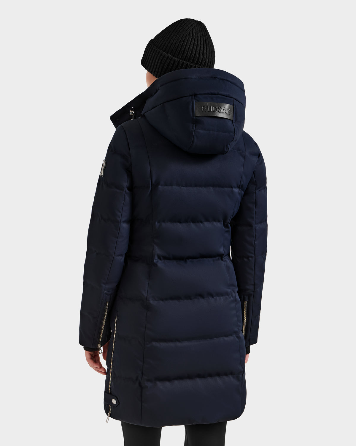 A person seen from the back wearing a long, navy Rudsak KEHLANI H - 8125643 down winter coat with a hood and a black knit beanie, set against a plain light background.